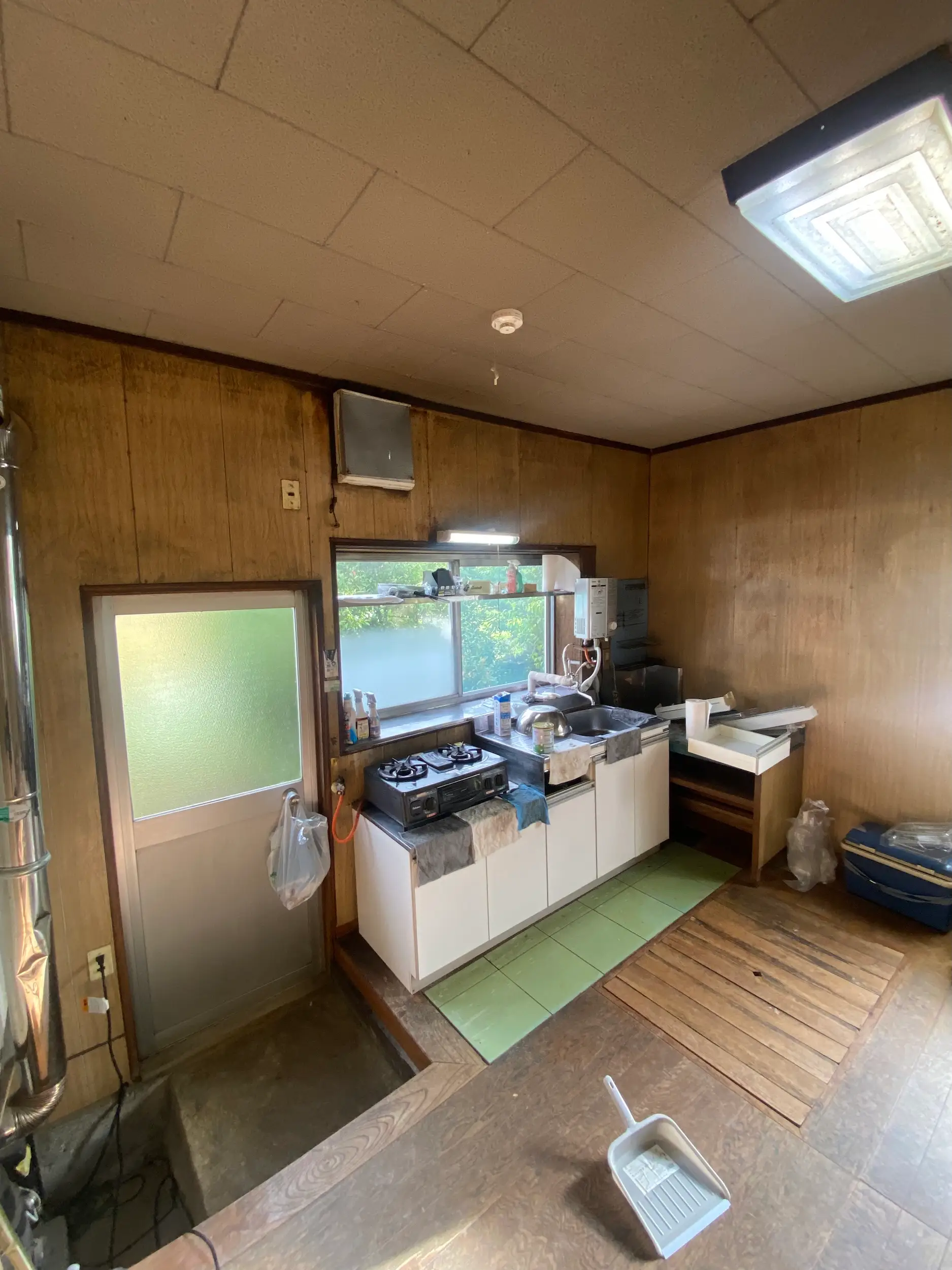 Small wood-paneled kitchen with a sink, gas stove, frosted door, window, and cleaning dustpan on the floor.