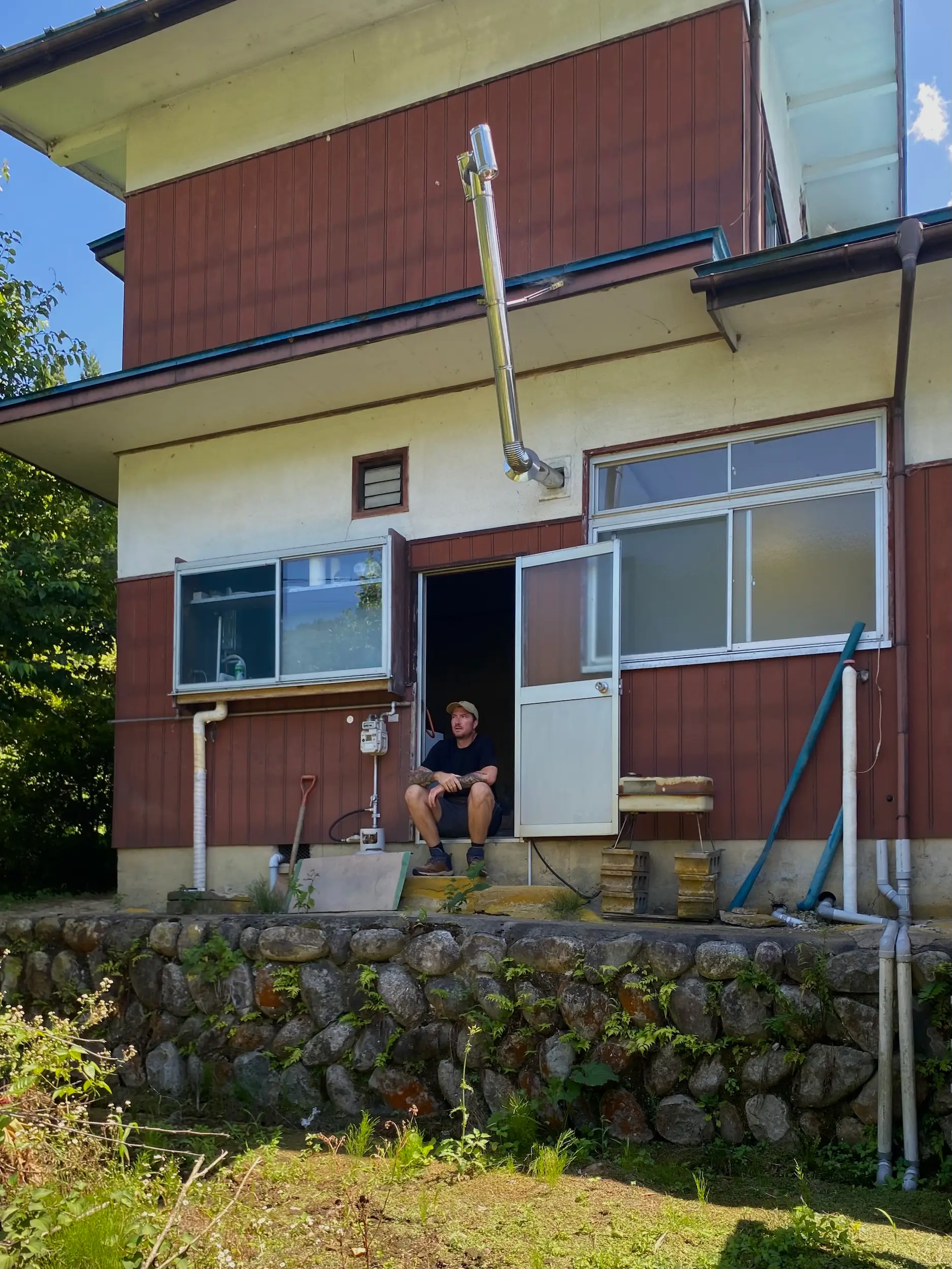 A person sits in an open doorway of a red and cream house with exterior pipes and stone foundation.