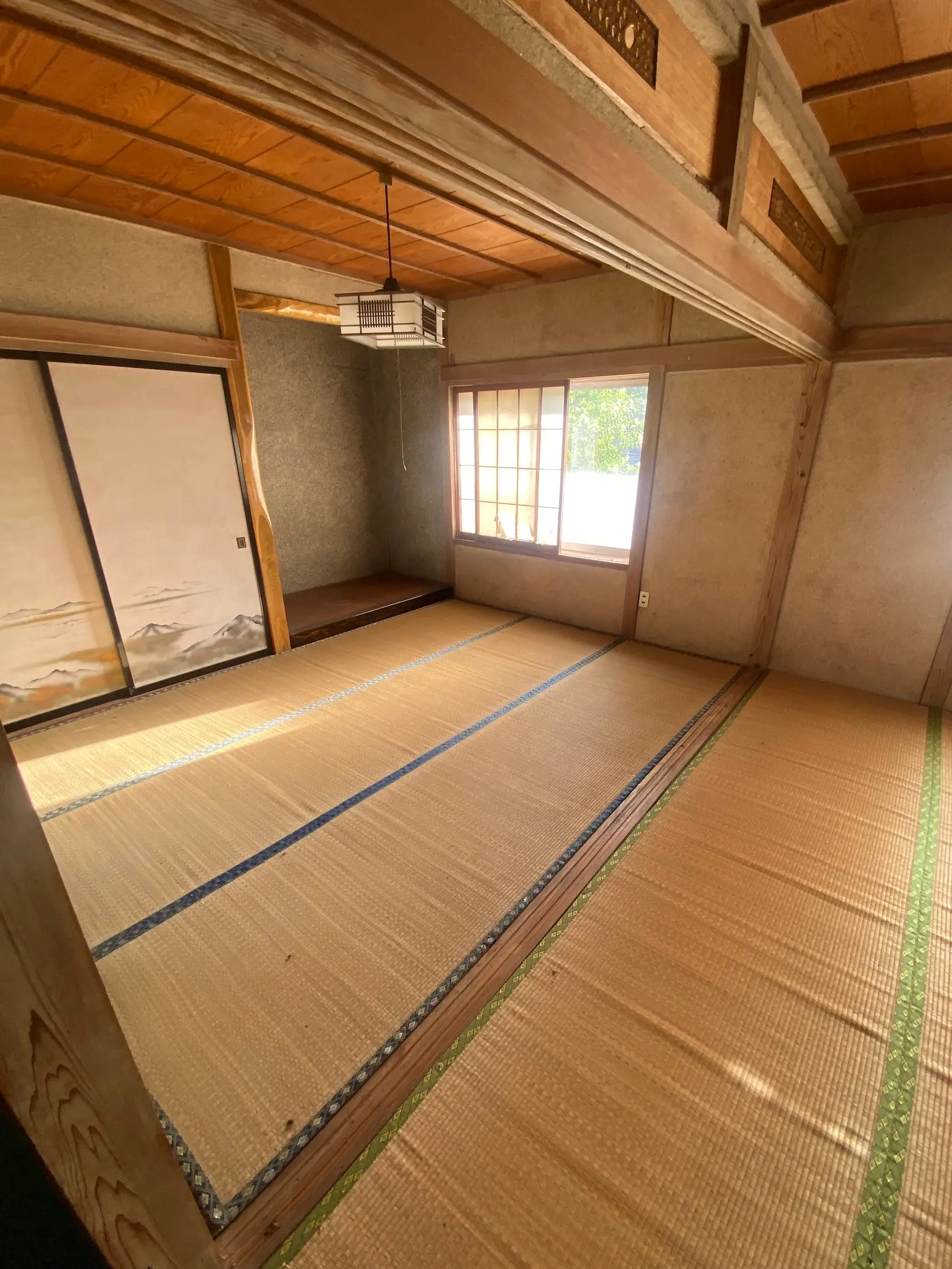 Empty Japanese-style tatami room with wooden beams, sliding doors, a window, and a hanging ceiling light.