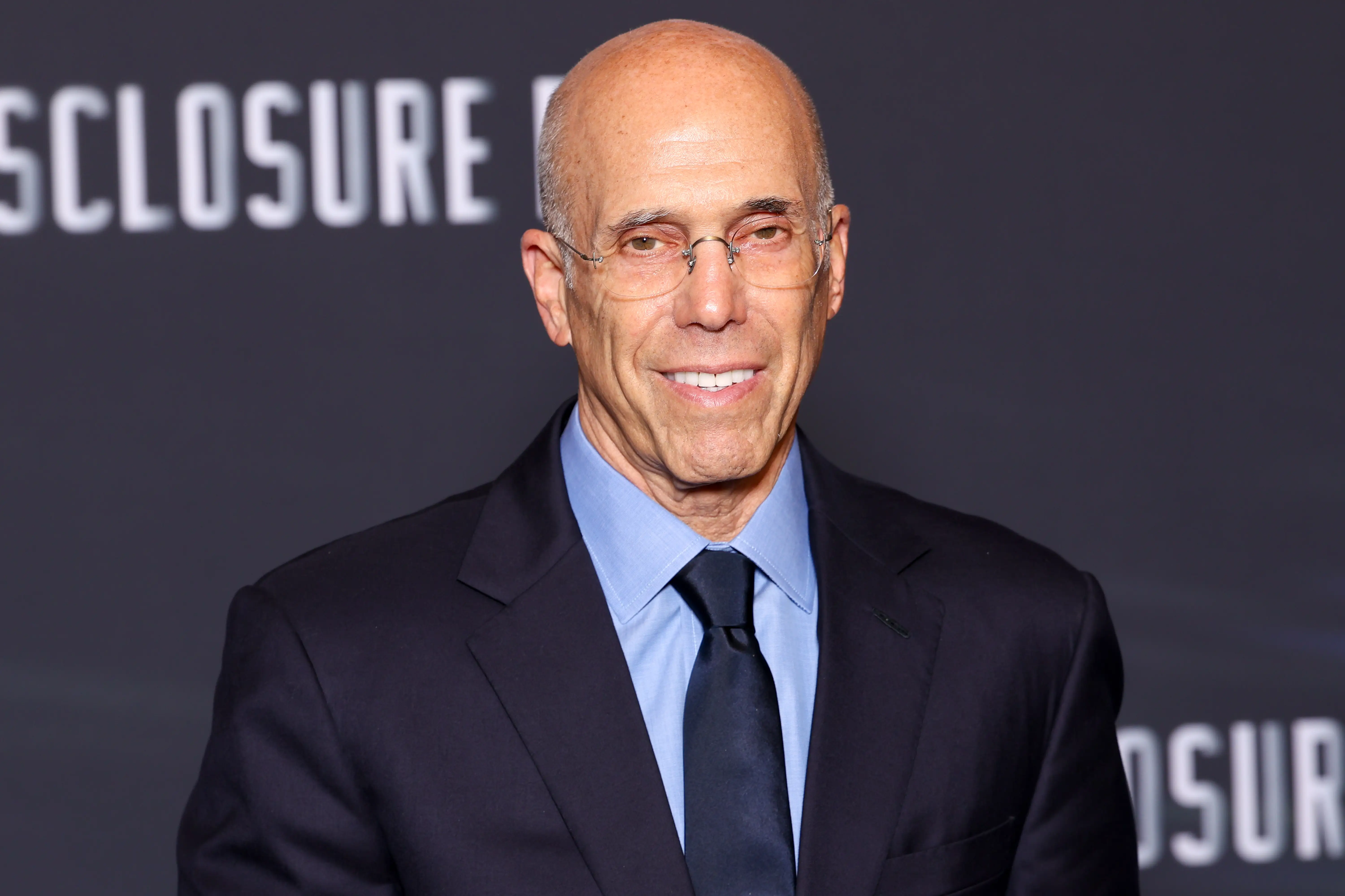 Jeffrey Katzenberg poses in a dark suit and glasses before a backdrop with partial 