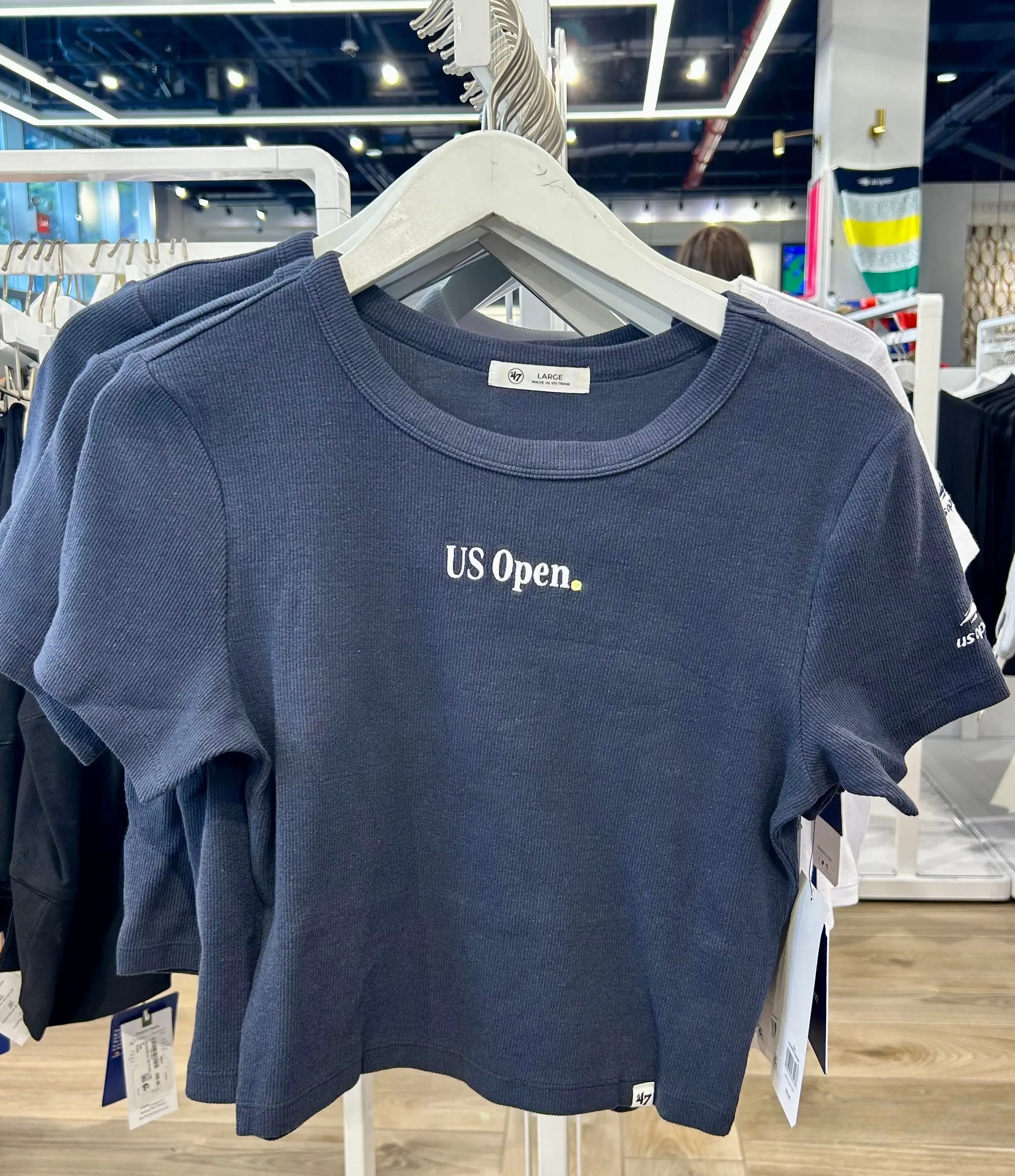 Navy blue US Open cropped shirt hangs on a white clothing rack inside a retail store.