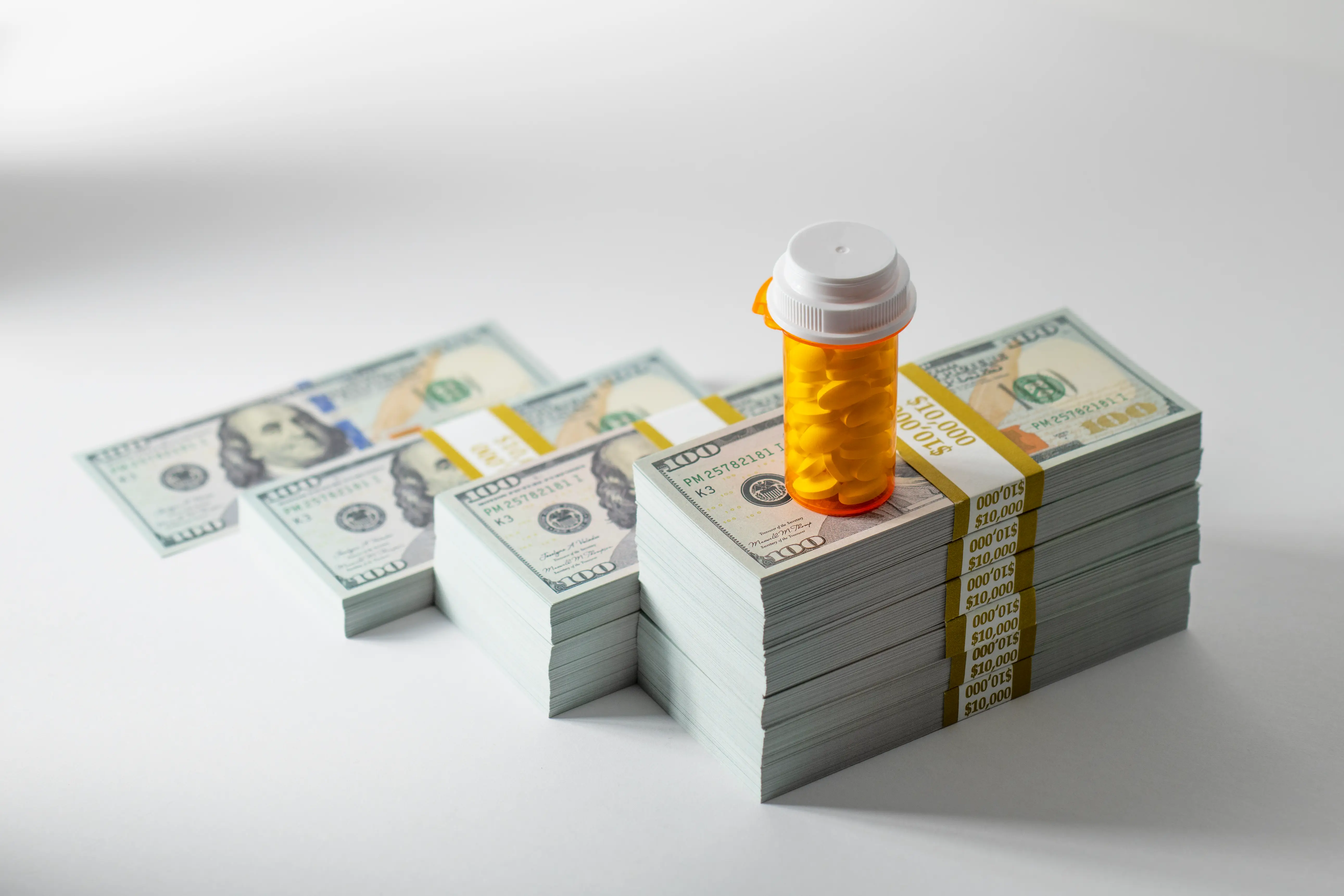 Prescription pill bottle with yellow tablets on growing stacks of US $100 bills, conceptual image representing pharmaceutical expenses, healthcare inflation, medication debt, and the financial burden of medication