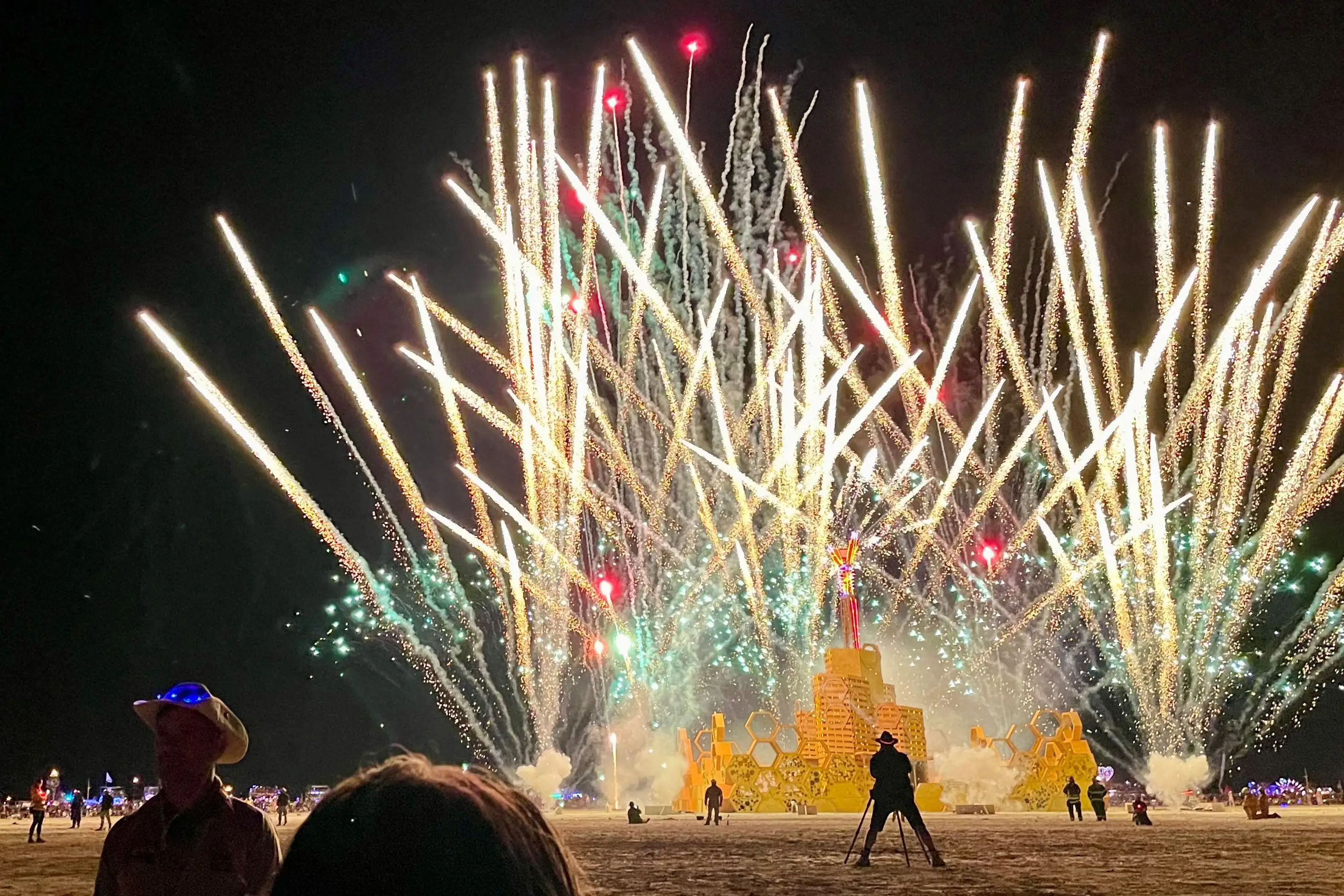 Fireworks explode over the Burning Man effigy as spectators watch from a dark desert playa.