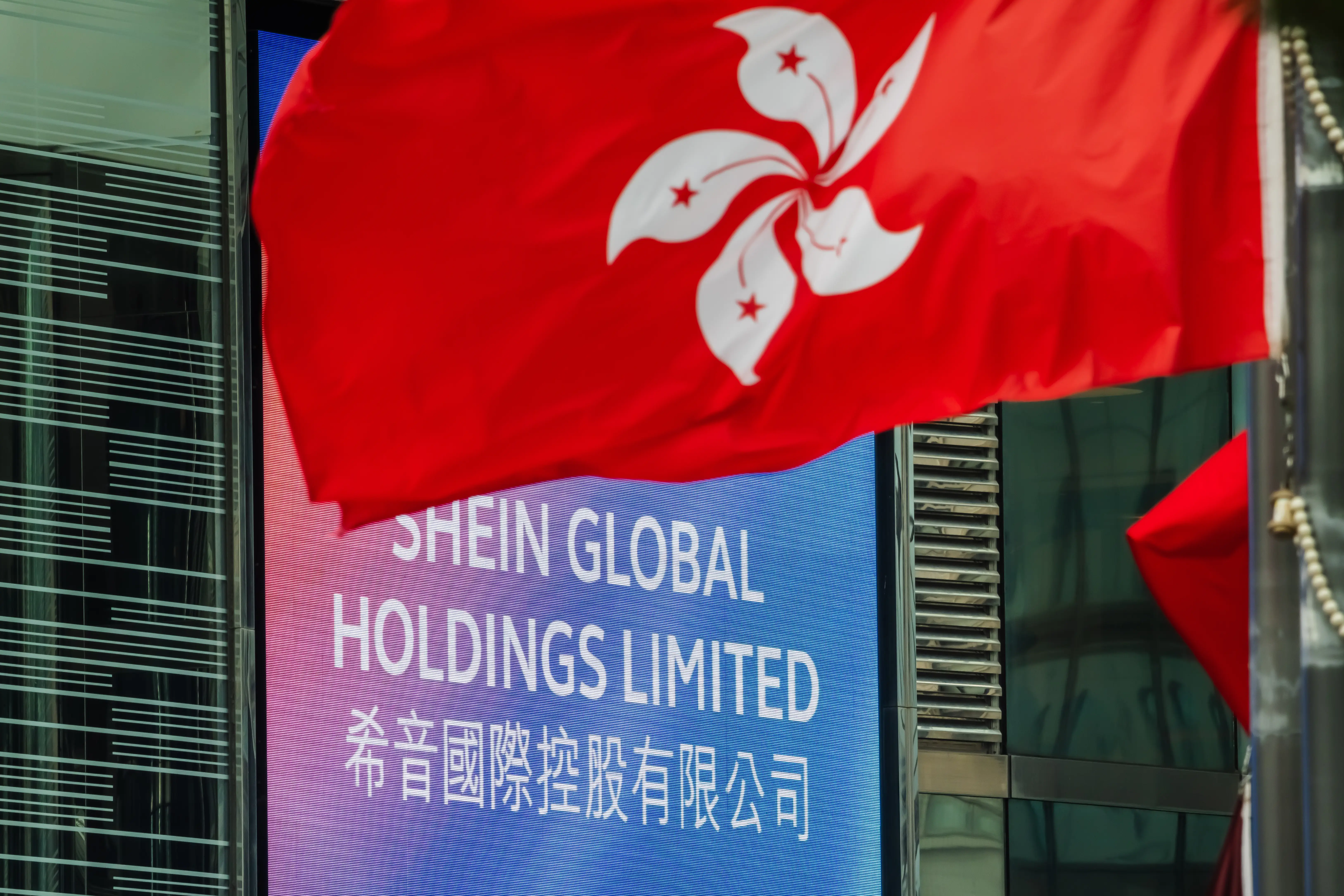 The Hong Kong flag flies in front of a large outdoor screen displaying the name of Shein Global Holdings Limited at Exchange Square, home to Hong Kong Exchanges and Clearing (HKEX), on September 1, 2026, in Hong Kong, China.