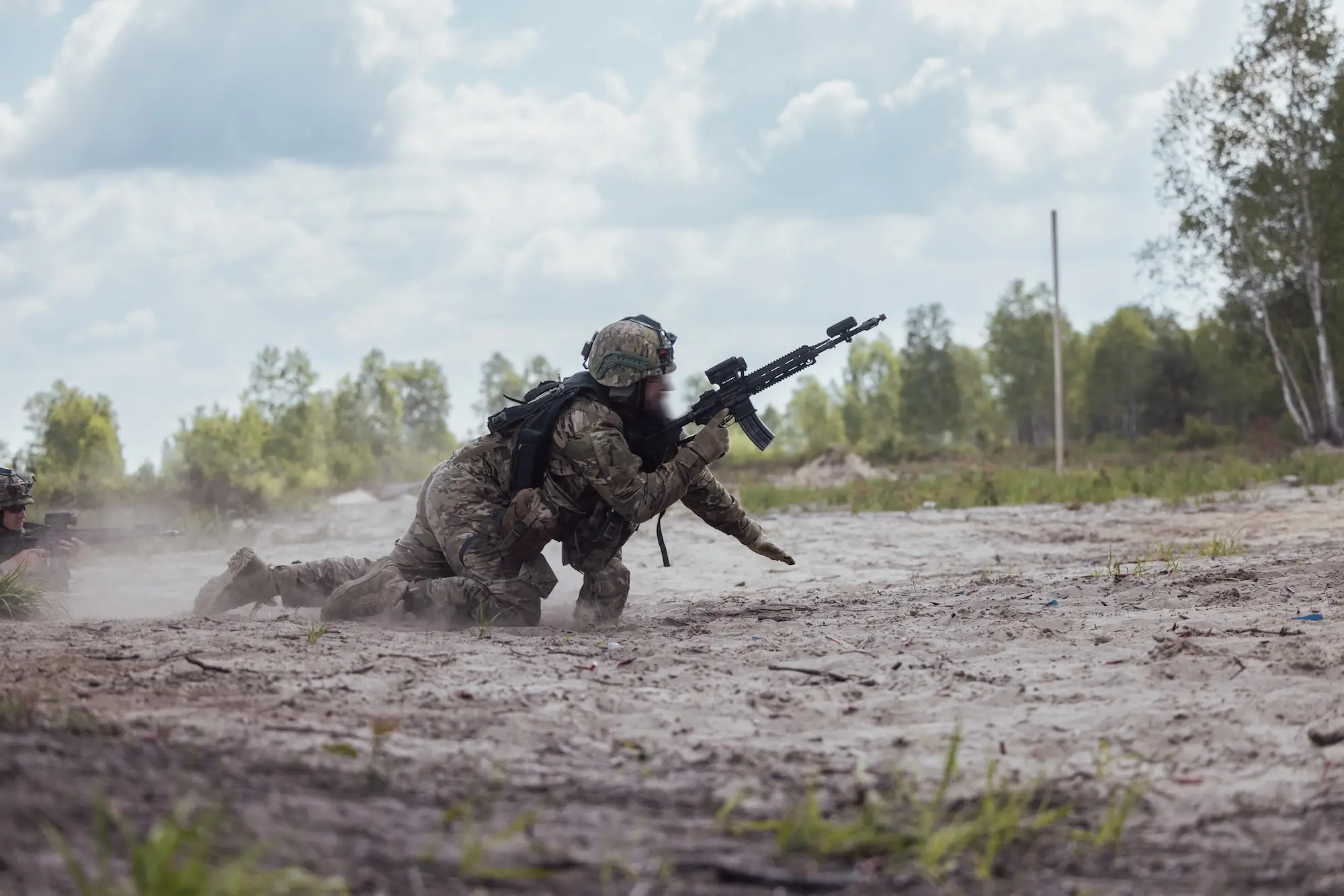 Camouflaged soldiers crawl through a dusty training field with rifles under a cloudy sky.
