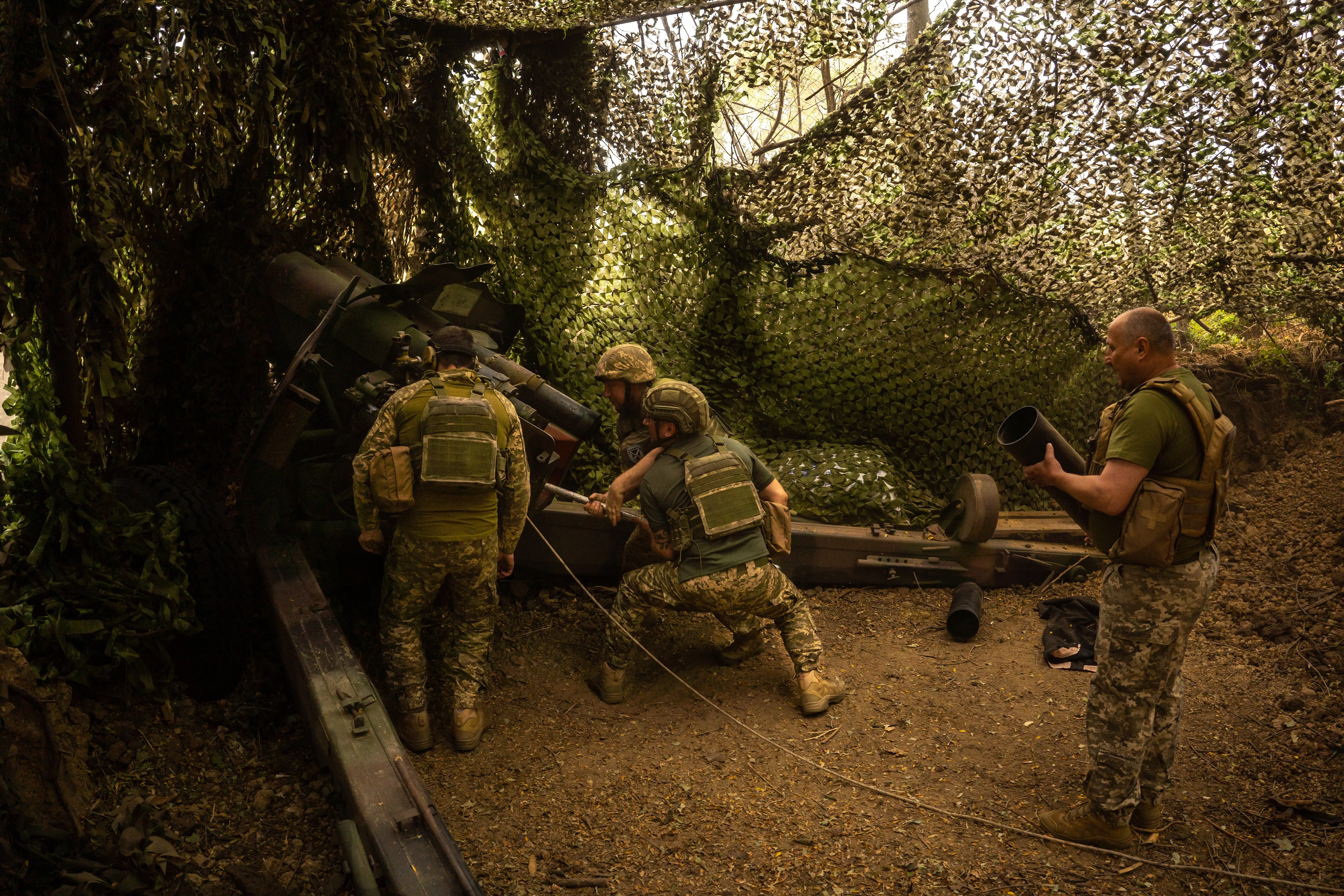 Service members operate a camouflaged artillery piece under netting in a wooded defensive position.