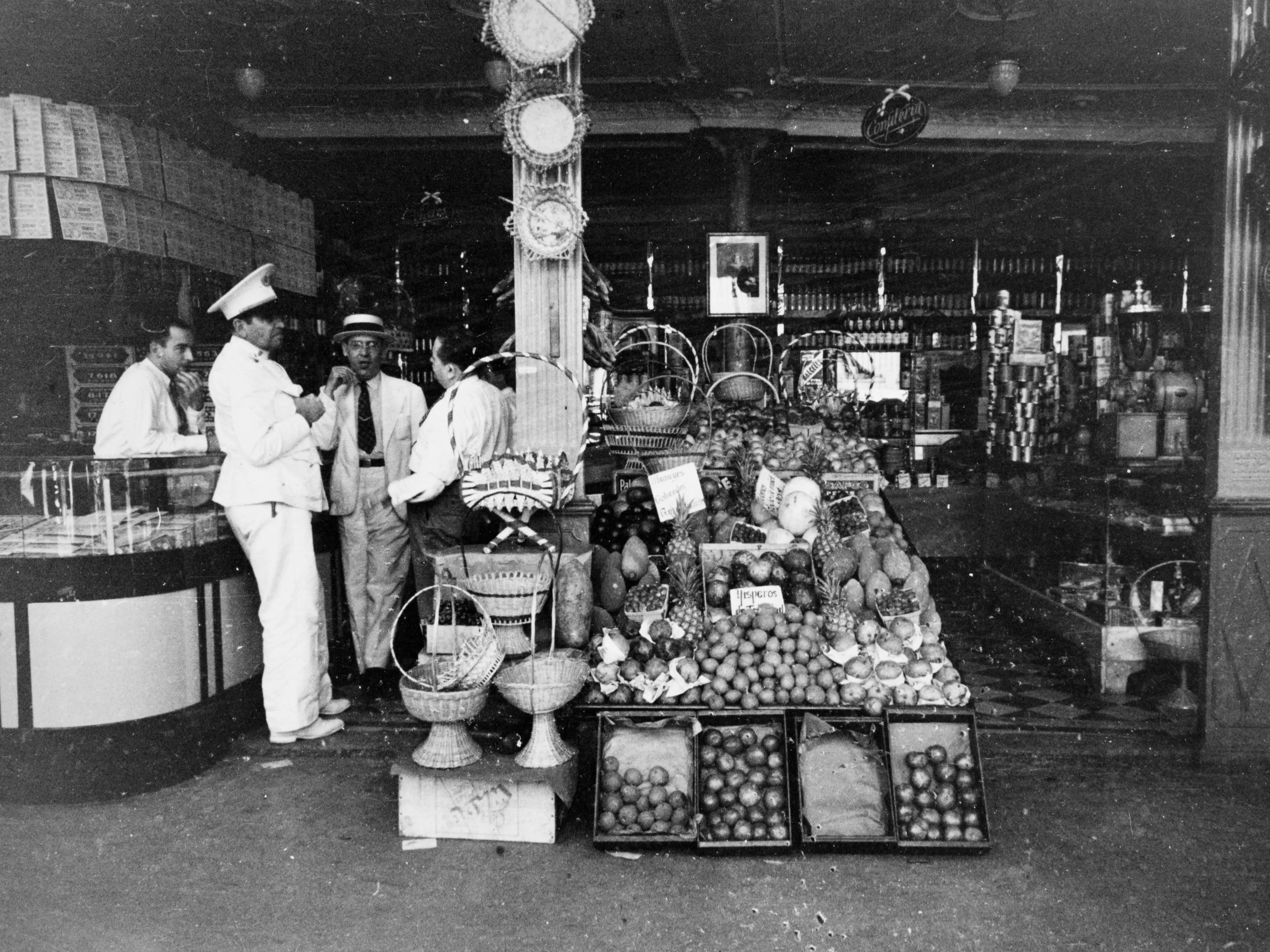 A group of men chatting near a shop counter inside a greengrocer and food shop, Havan, Cuba, circa 1950.
