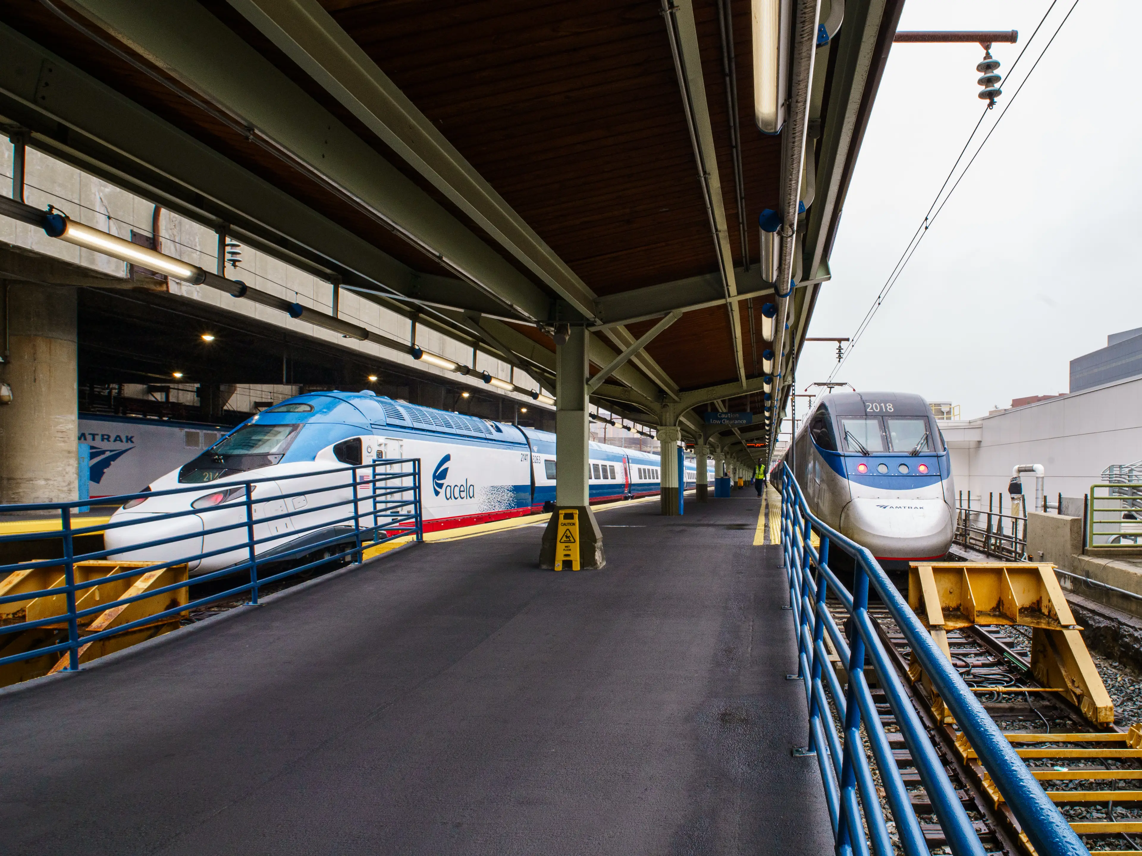 Two Amtrak Acela trains stand on opposite tracks beside a covered station platform with blue railings.