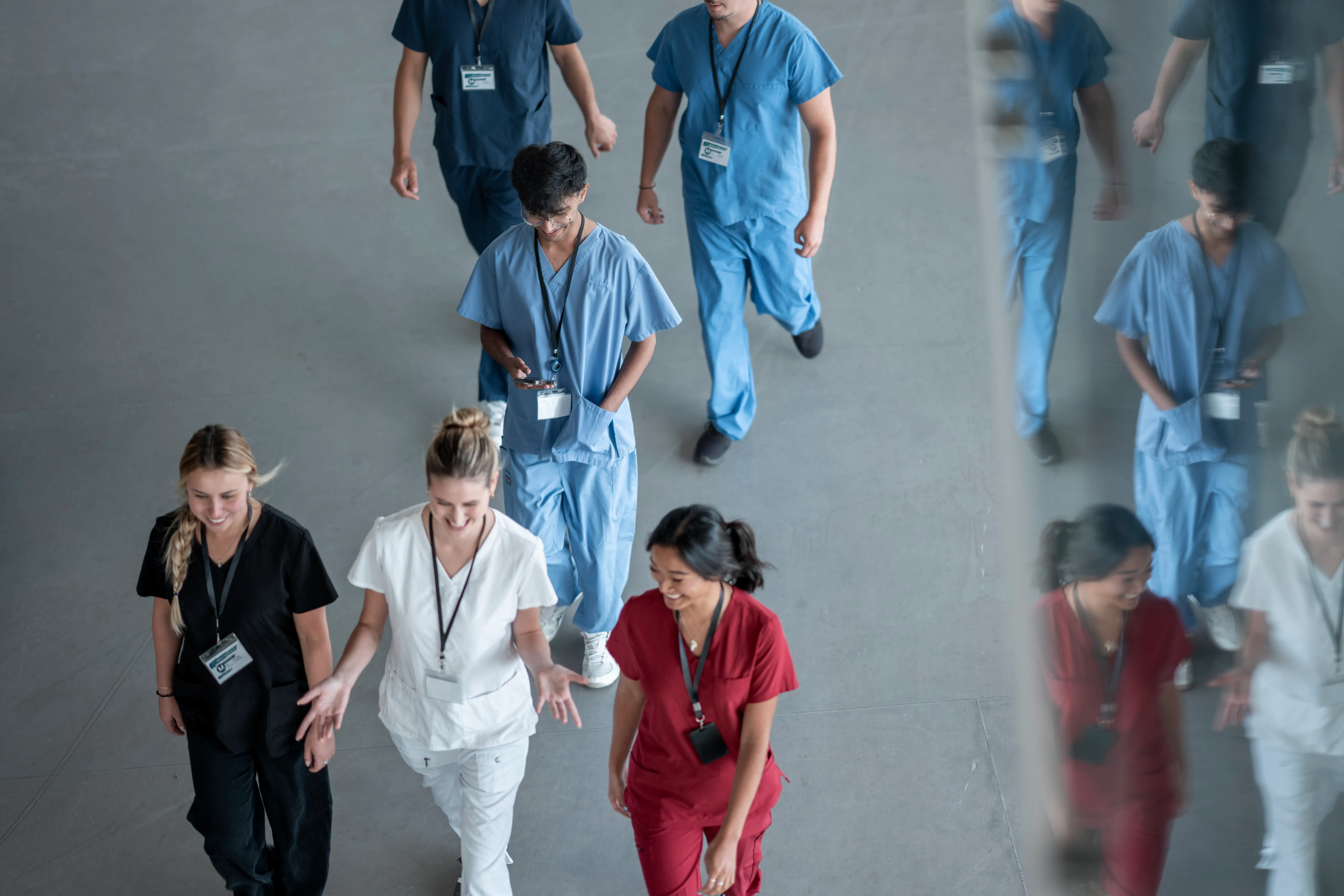 Group of people in medical scrubs with ID badges walking through a corridor beside a reflective wall.