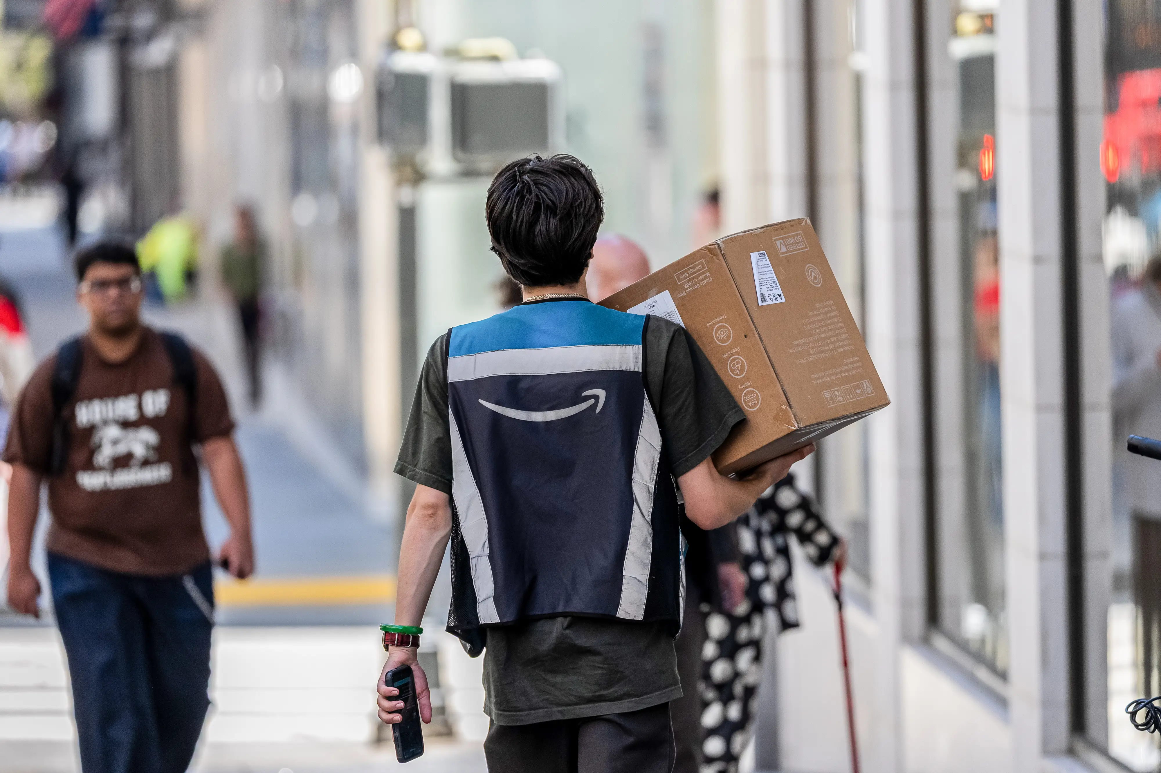 An Amazon delivery worker carries a package in one hand and a phone in the other as they wear an Amazon-branded reflective vest.