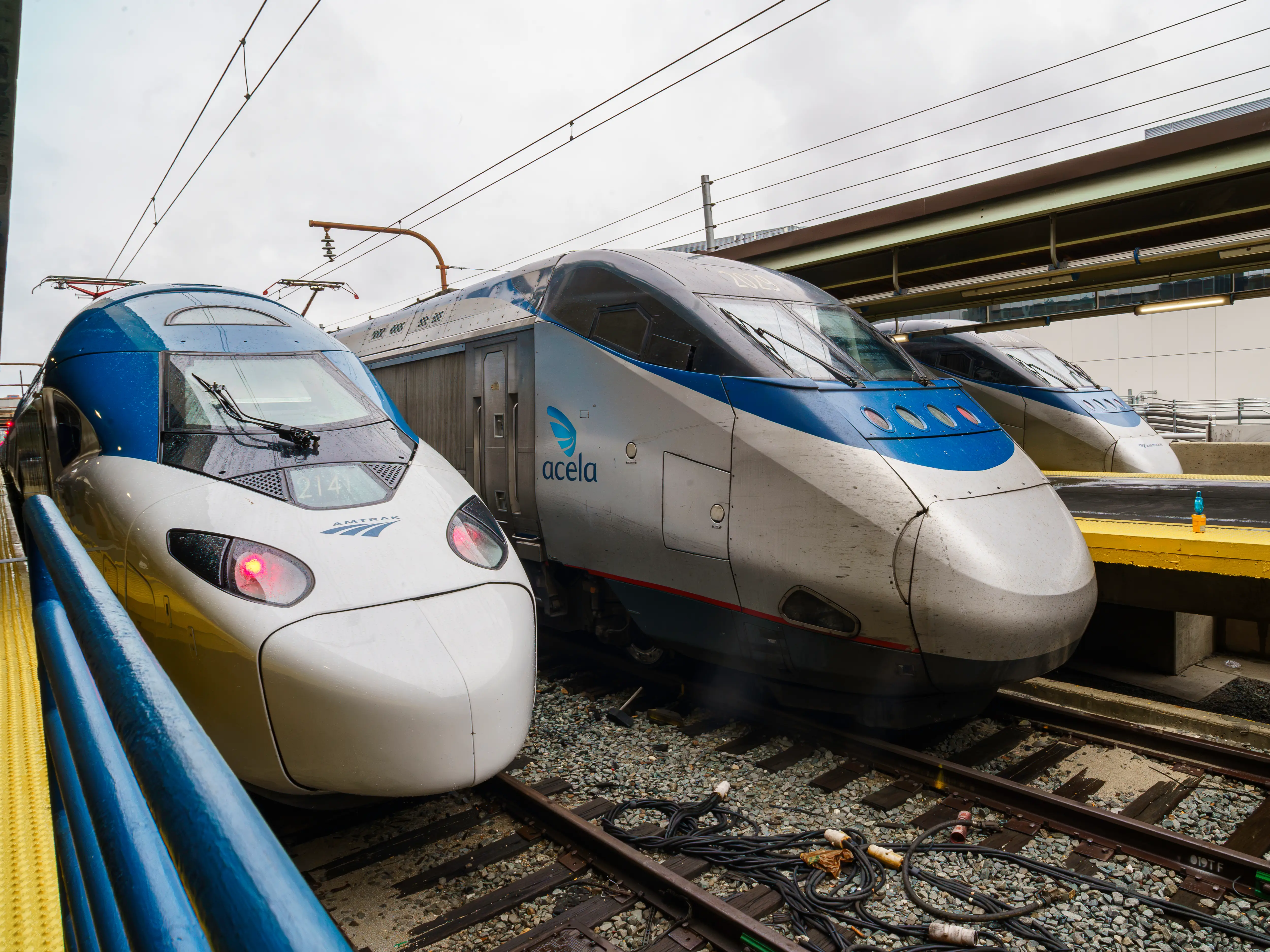 Three Amtrak Acela trains stand side by side on wet tracks at an overcast station platform.