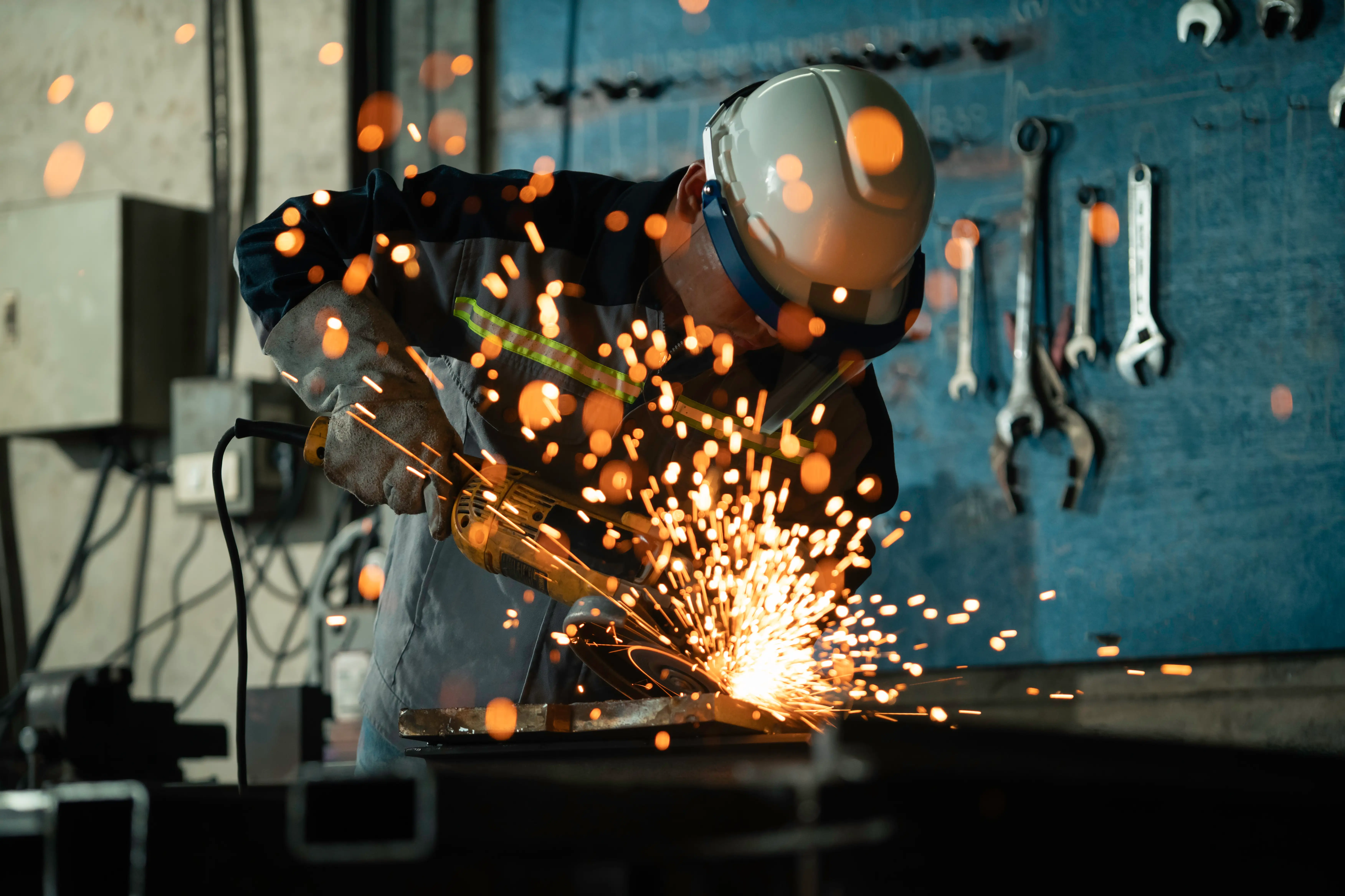 Worker wearing protective gear uses an angle grinder on metal as bright sparks fly in a workshop.