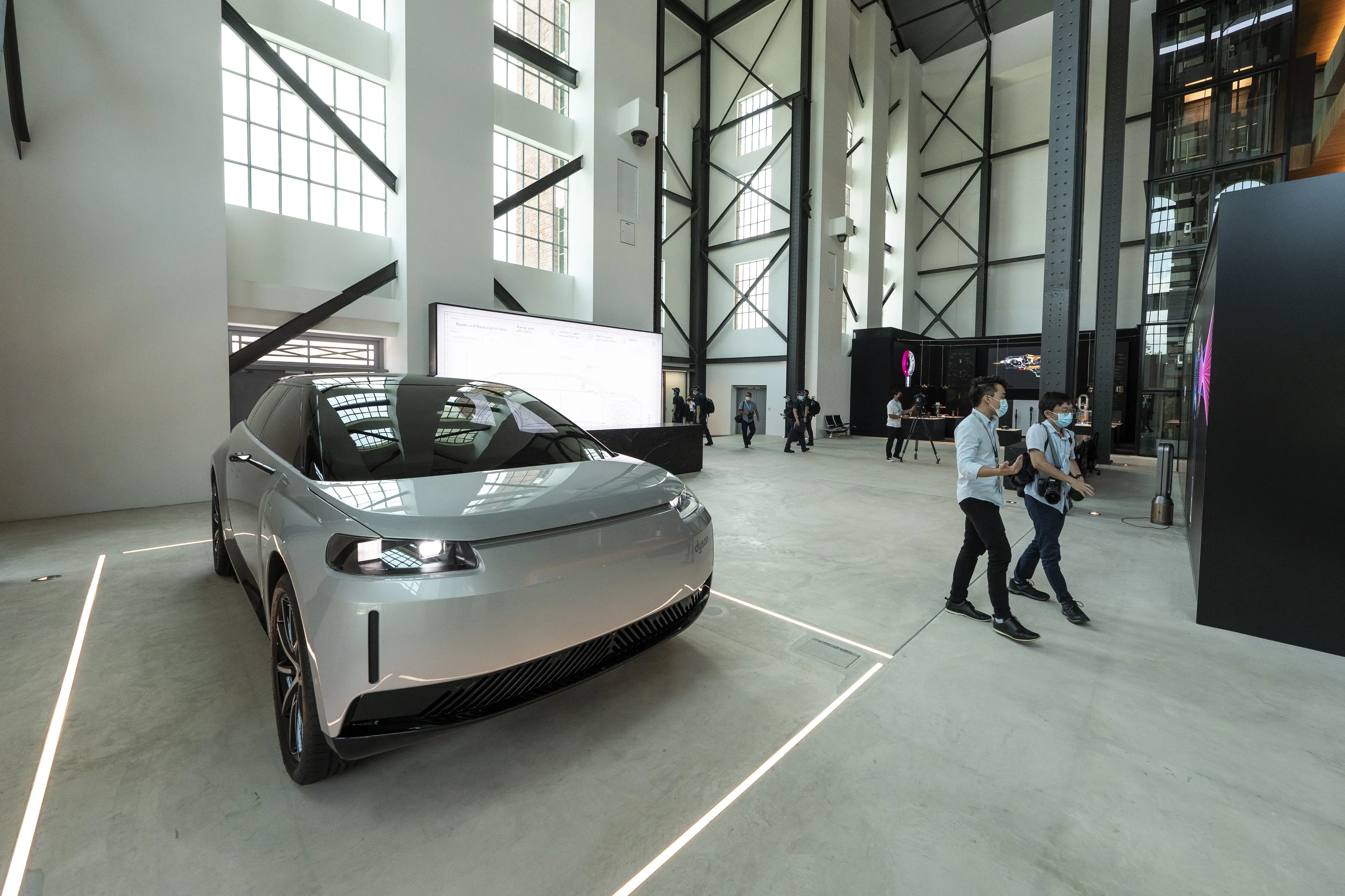 A Silver Dyson EV prototype sits in a warehouse room. A few people dressed in semi-formal clothes walk around.