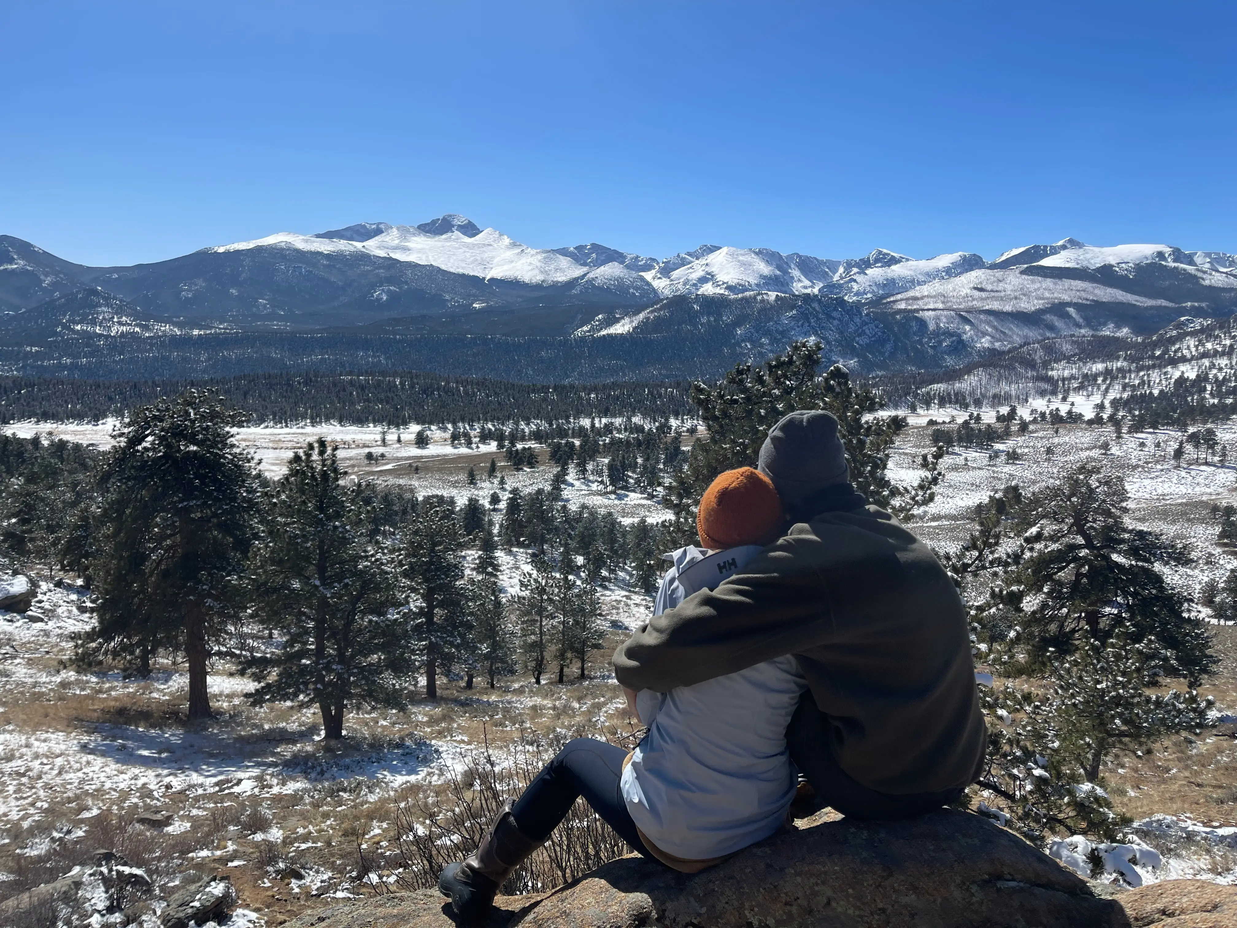 Two people sitting on mountain looking at mountains, trees in distance