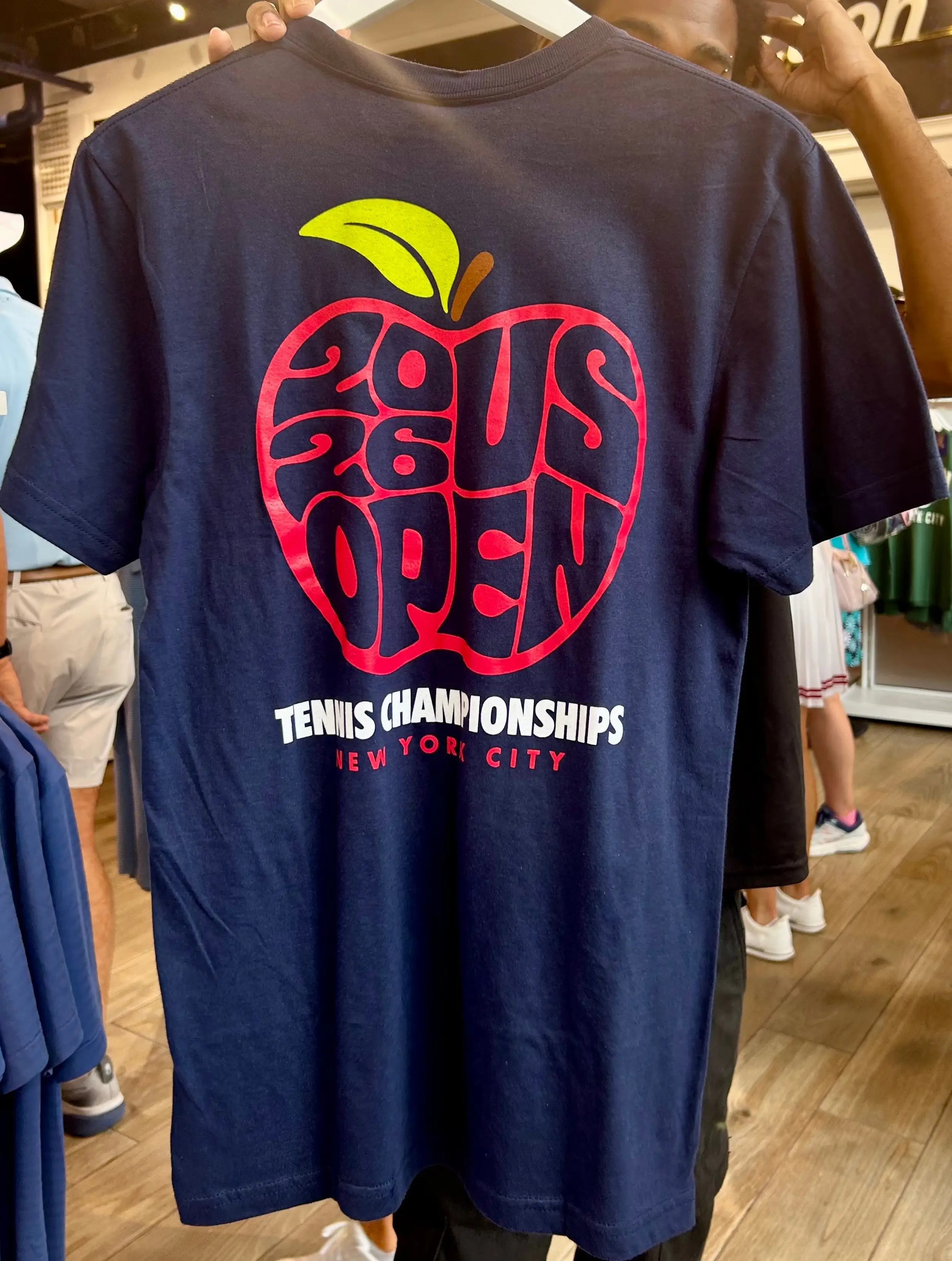 Navy T-shirt on a hanger displays a stylized apple graphic for 2025 US Open Tennis Championships in New York City.