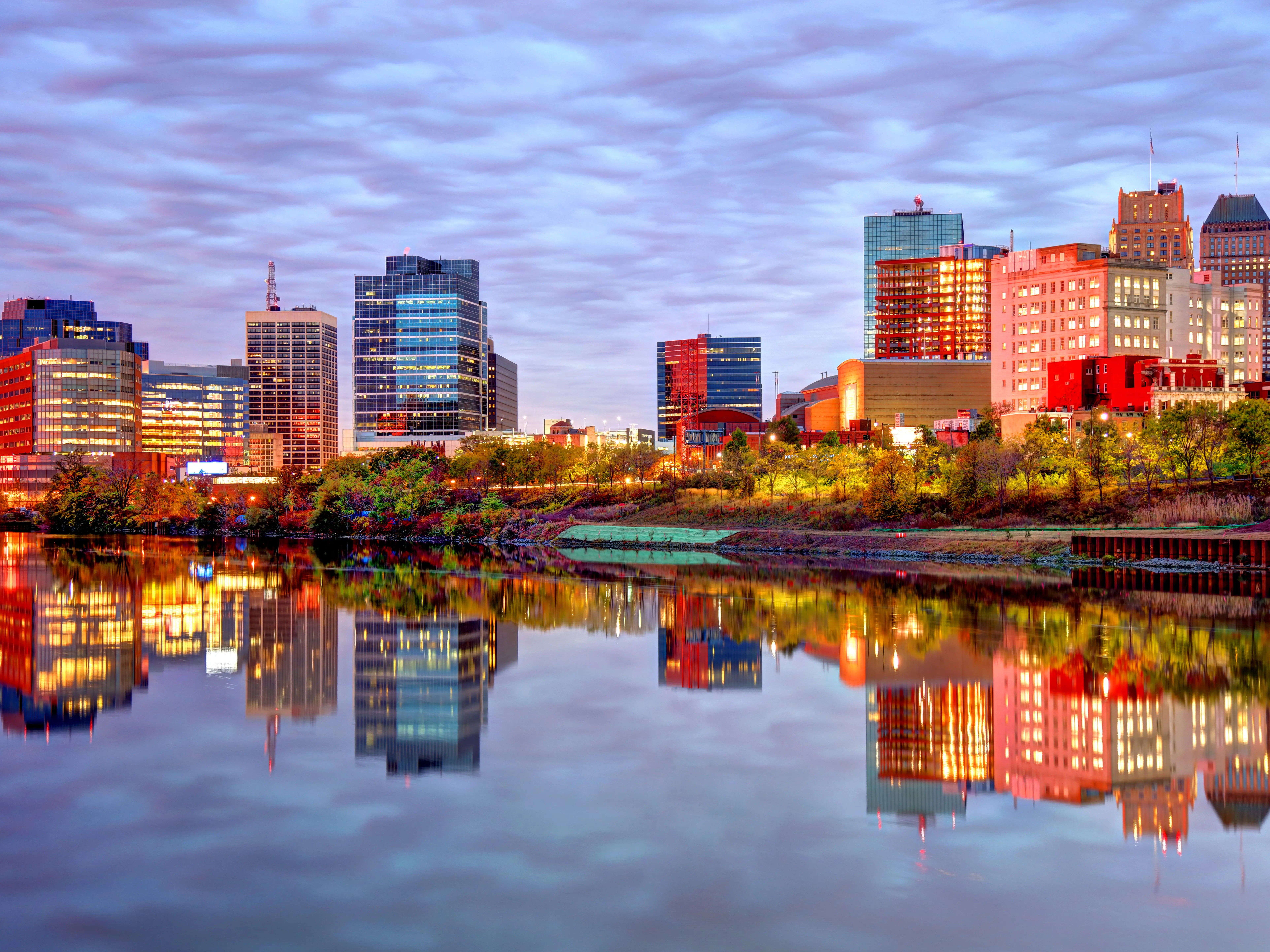 Illuminated downtown skyline reflects across a calm river at dusk under a cloud-filled sky.