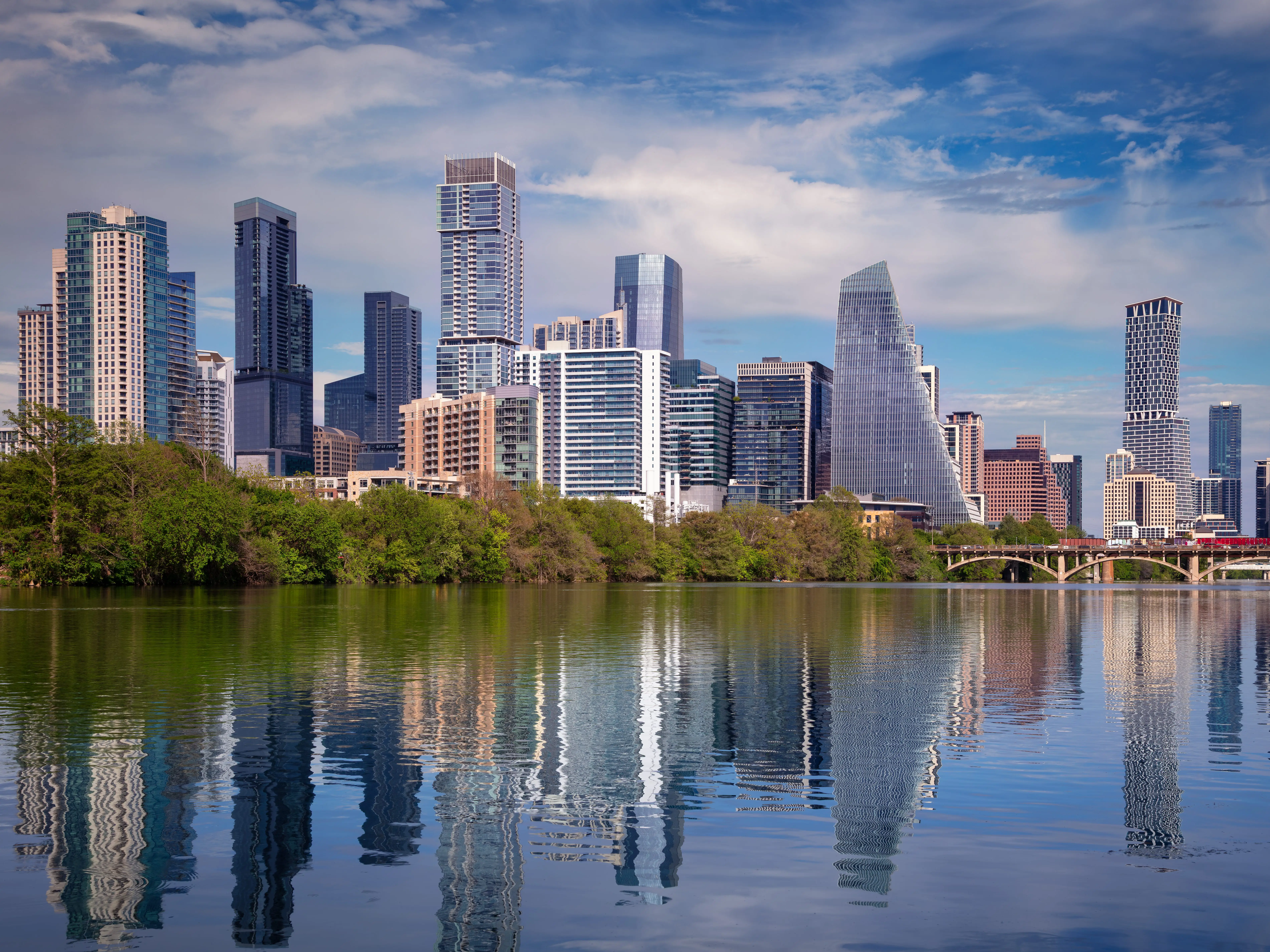 Austin, Texas skyline with modern skyscrapers and unique architectural designs against a blue sky. The foreground features a calm body of water reflecting the buildings and surrounding green trees. The scene captures a mix of urban architecture and natural beauty, showcasing Austin's vibrant cityscape and lush environment.