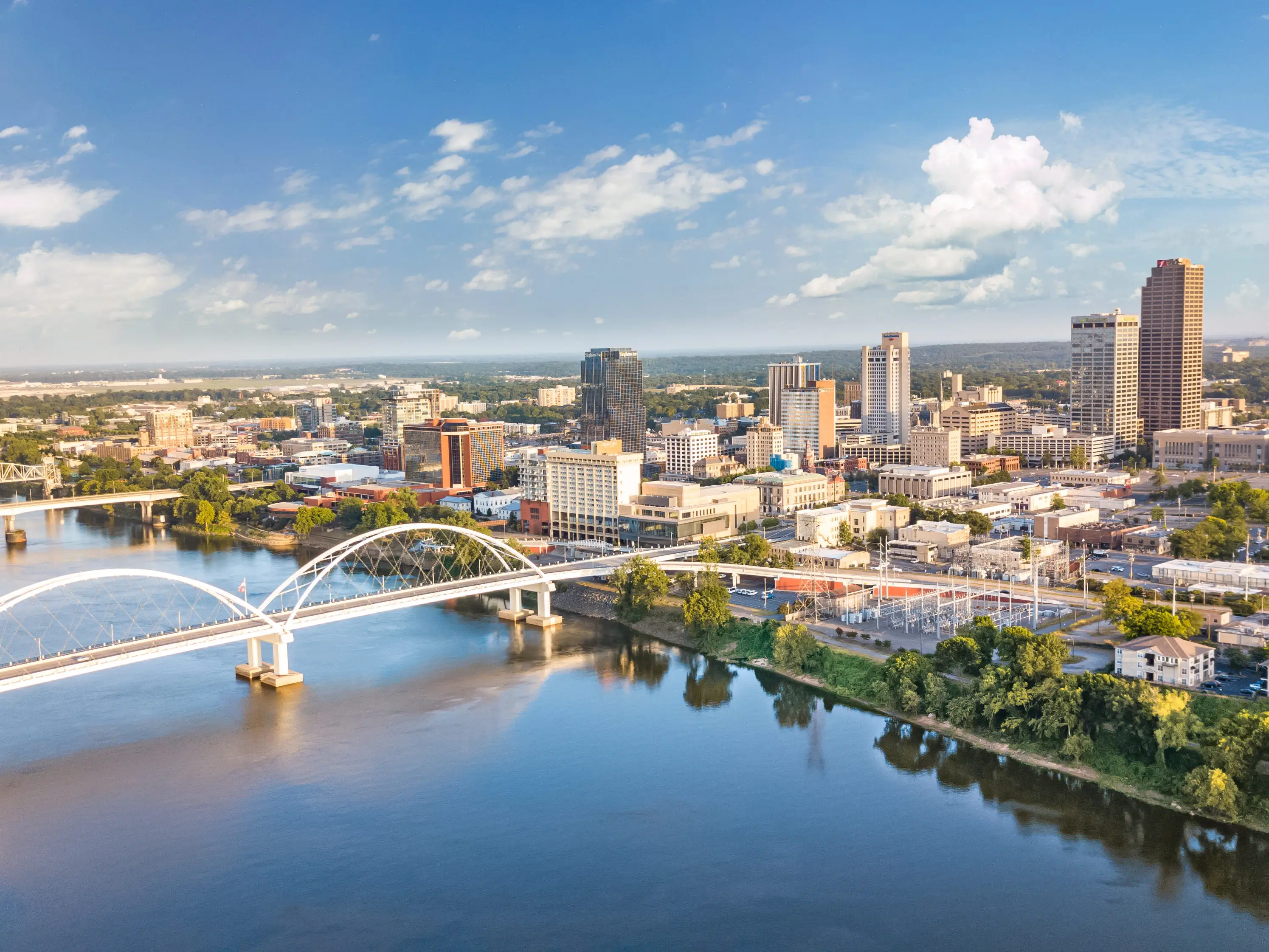 Downtown Little Rock skyline and a white arch bridge span the Arkansas River under a blue sky.