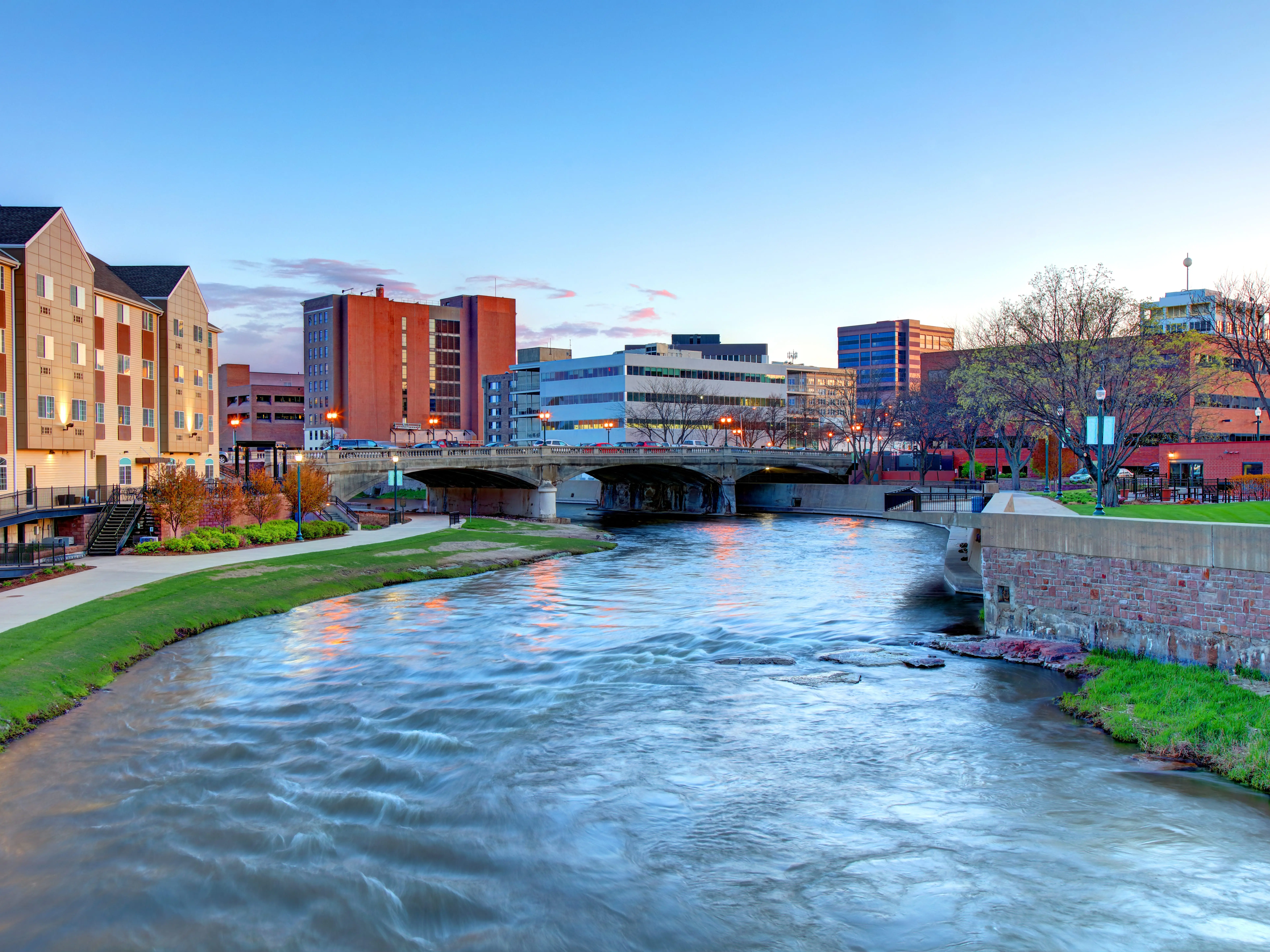 Urban river flows beneath a bridge between downtown buildings at dusk under a clear blue sky.