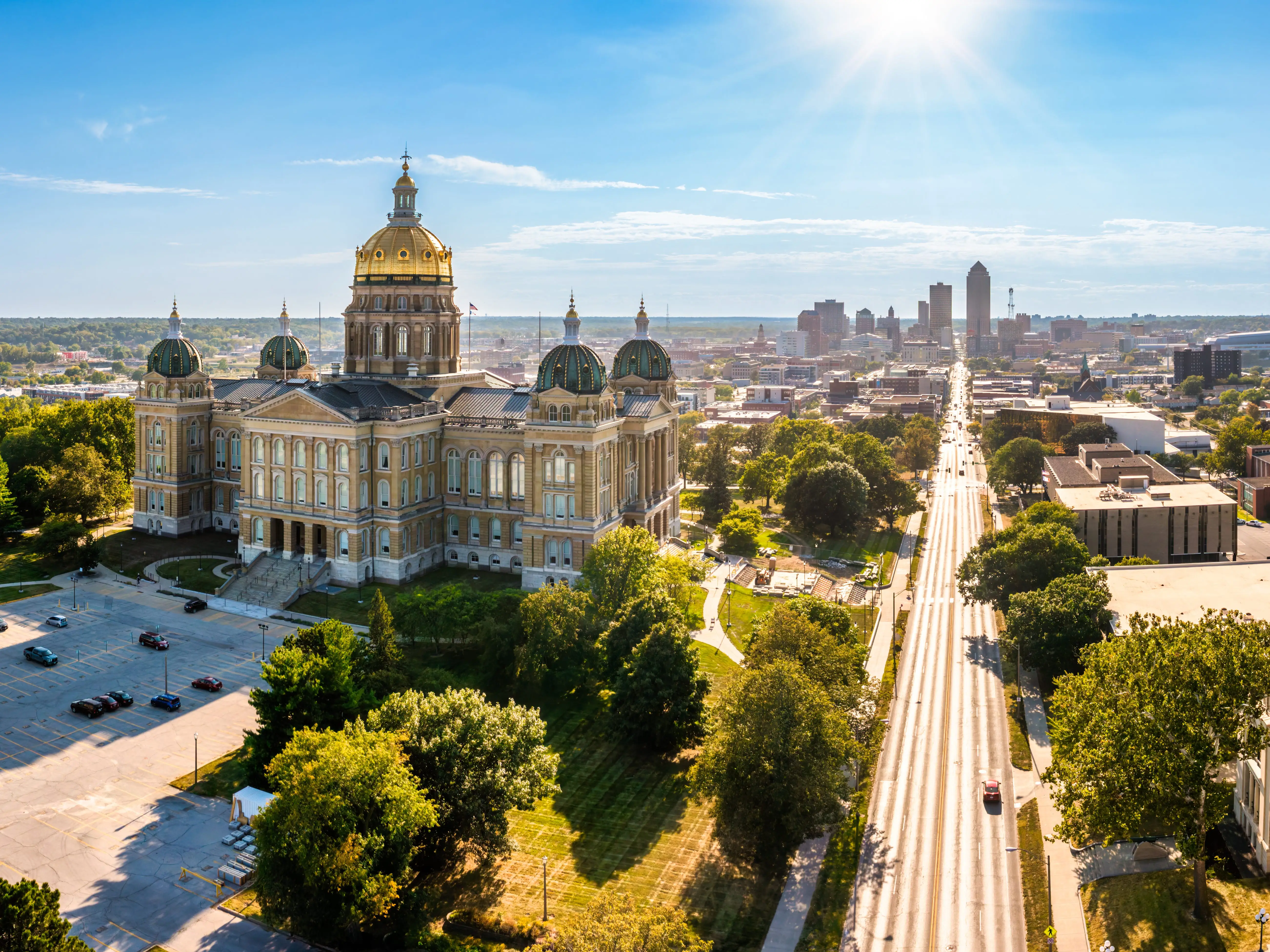Iowa State Capitol with gold dome overlooks a broad avenue and downtown Des Moines under bright sun.