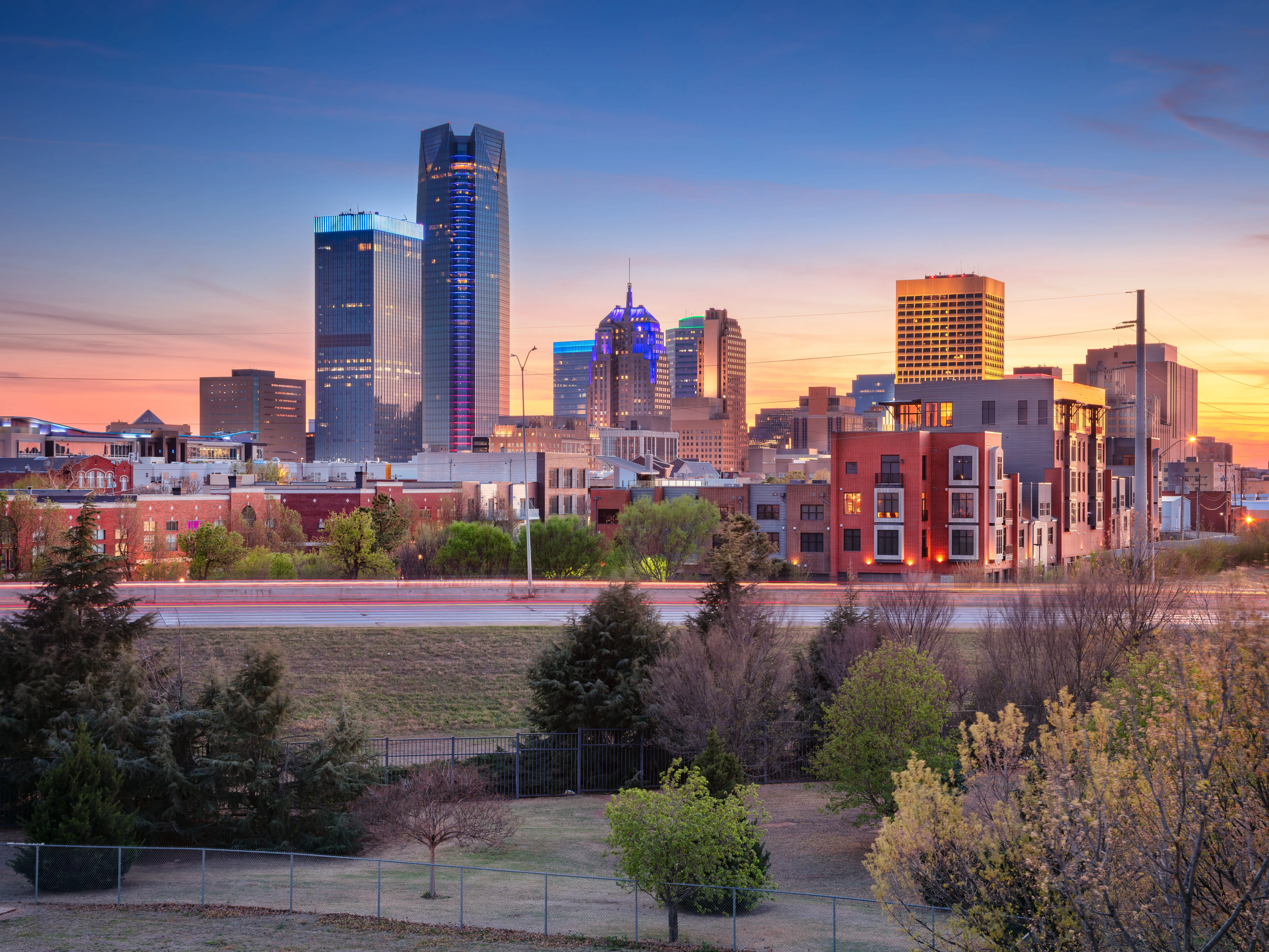 Downtown Oklahoma City skyline at sunset with high-rise towers, urban buildings, trees, and light trails in the foreground.