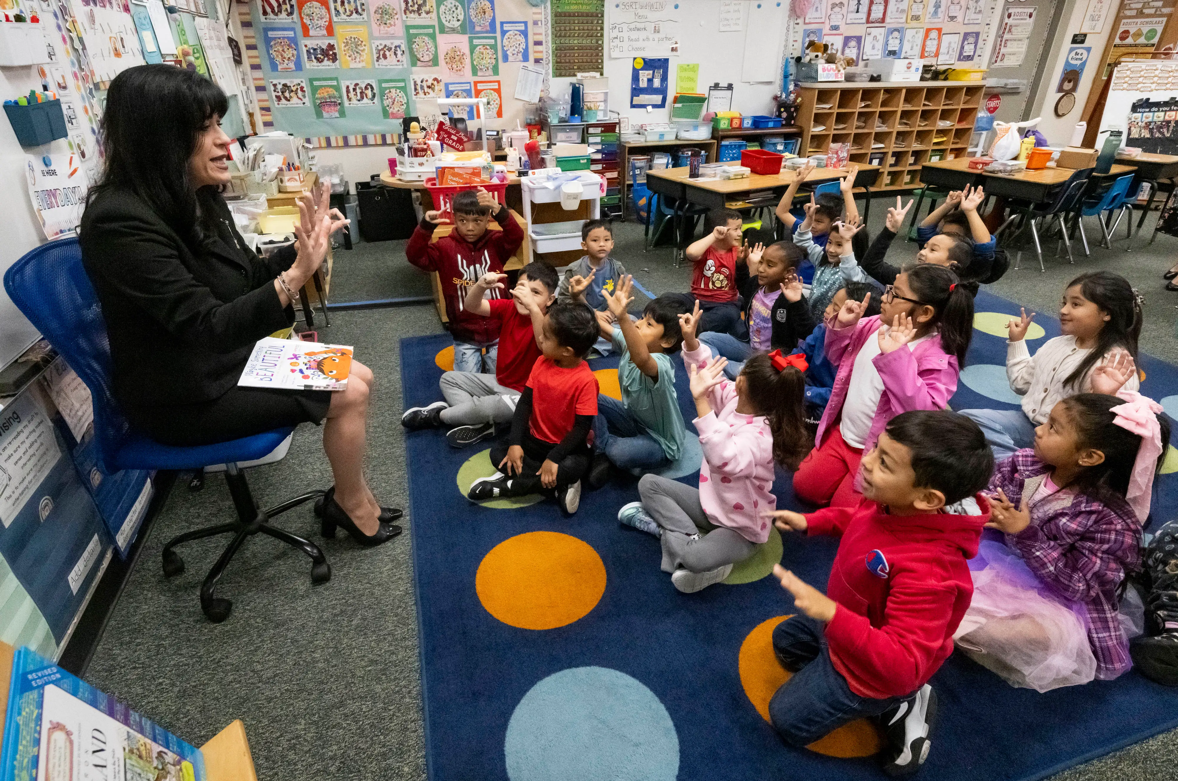 A group of small children sit on the ground in front of a teacher in a seat.