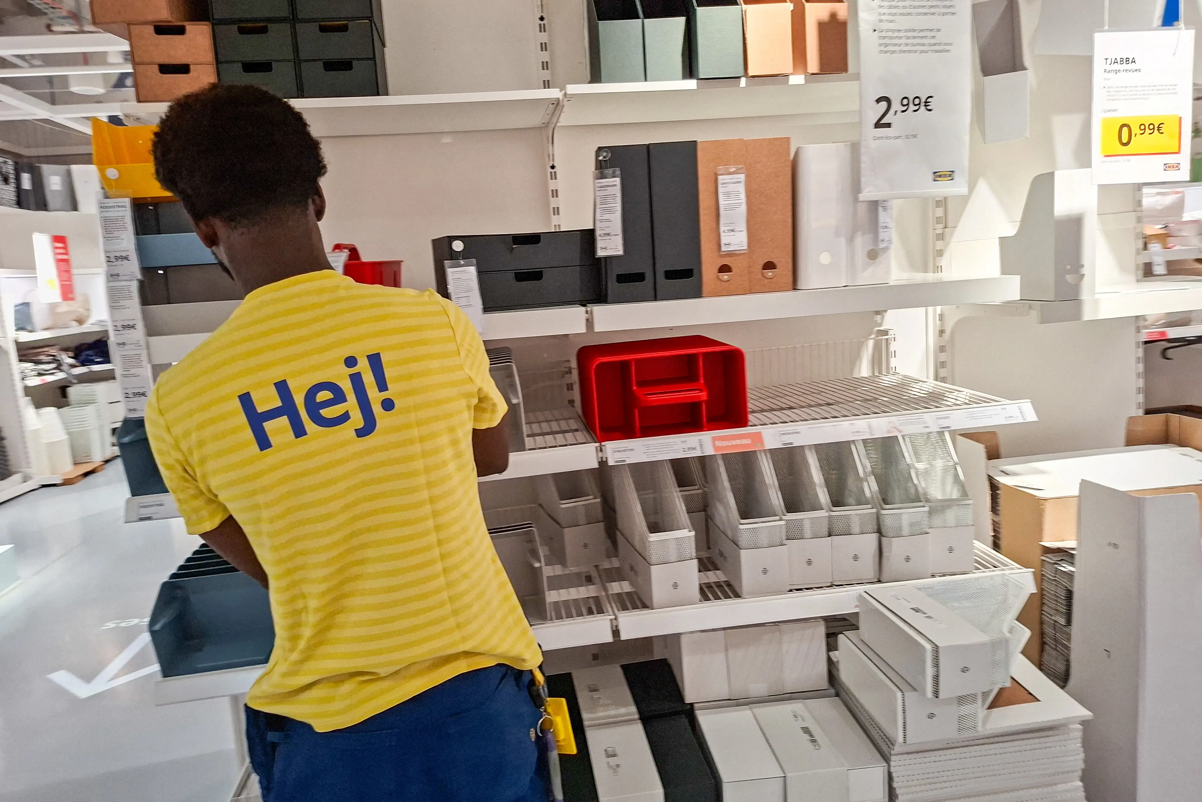 An employee wearing a yellow T-shirt with Hej printed on it stands in front of a storage aisle at the IKEA store in the Italie Deux shopping center in Paris, France, on July 27, 2026. Boxes, organizers, and magazine holders in various colors are displayed on the shelves, with a price tag of 2.99.
