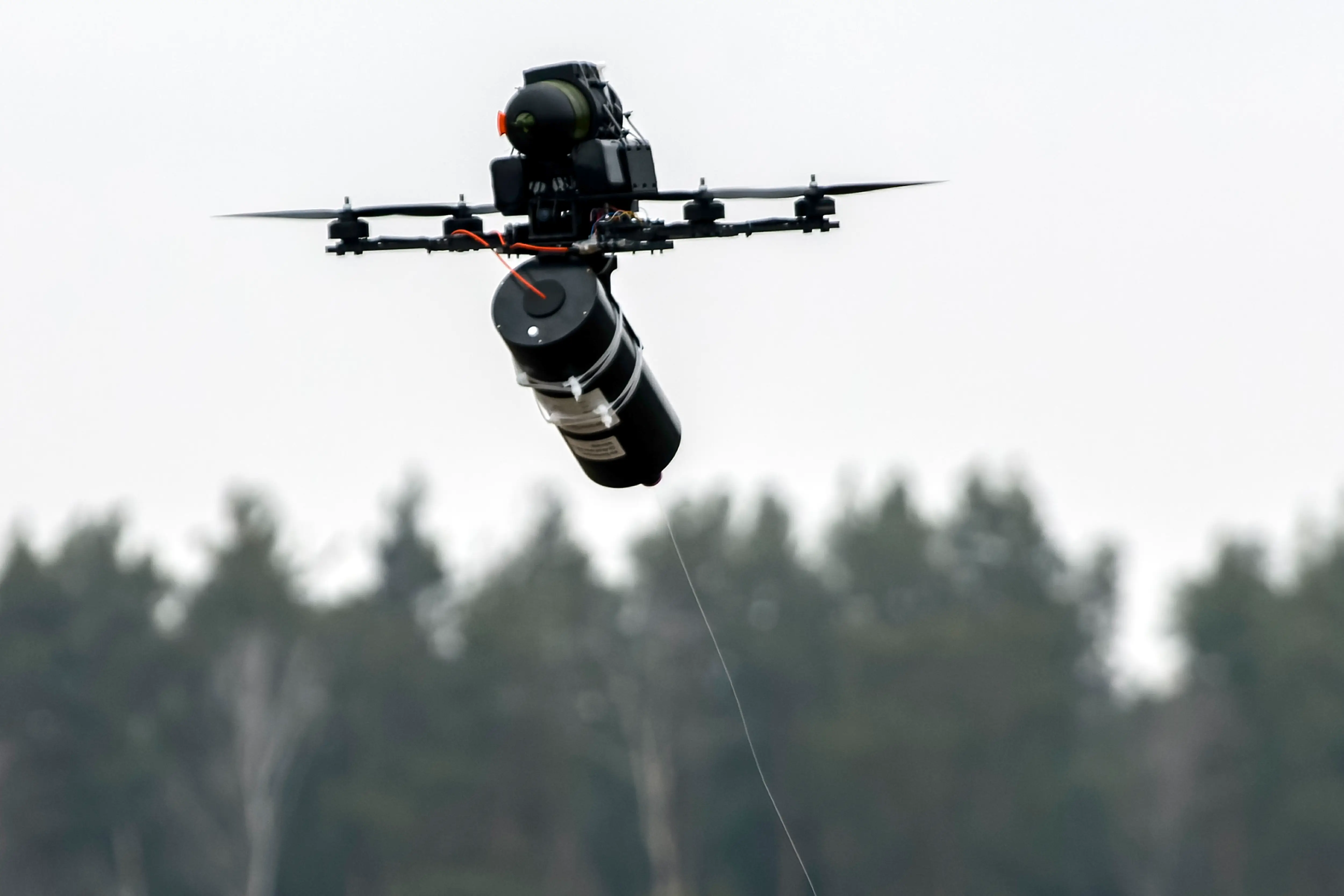 Black multi-rotor drone flies above blurred trees carrying a large suspended cylindrical payload.