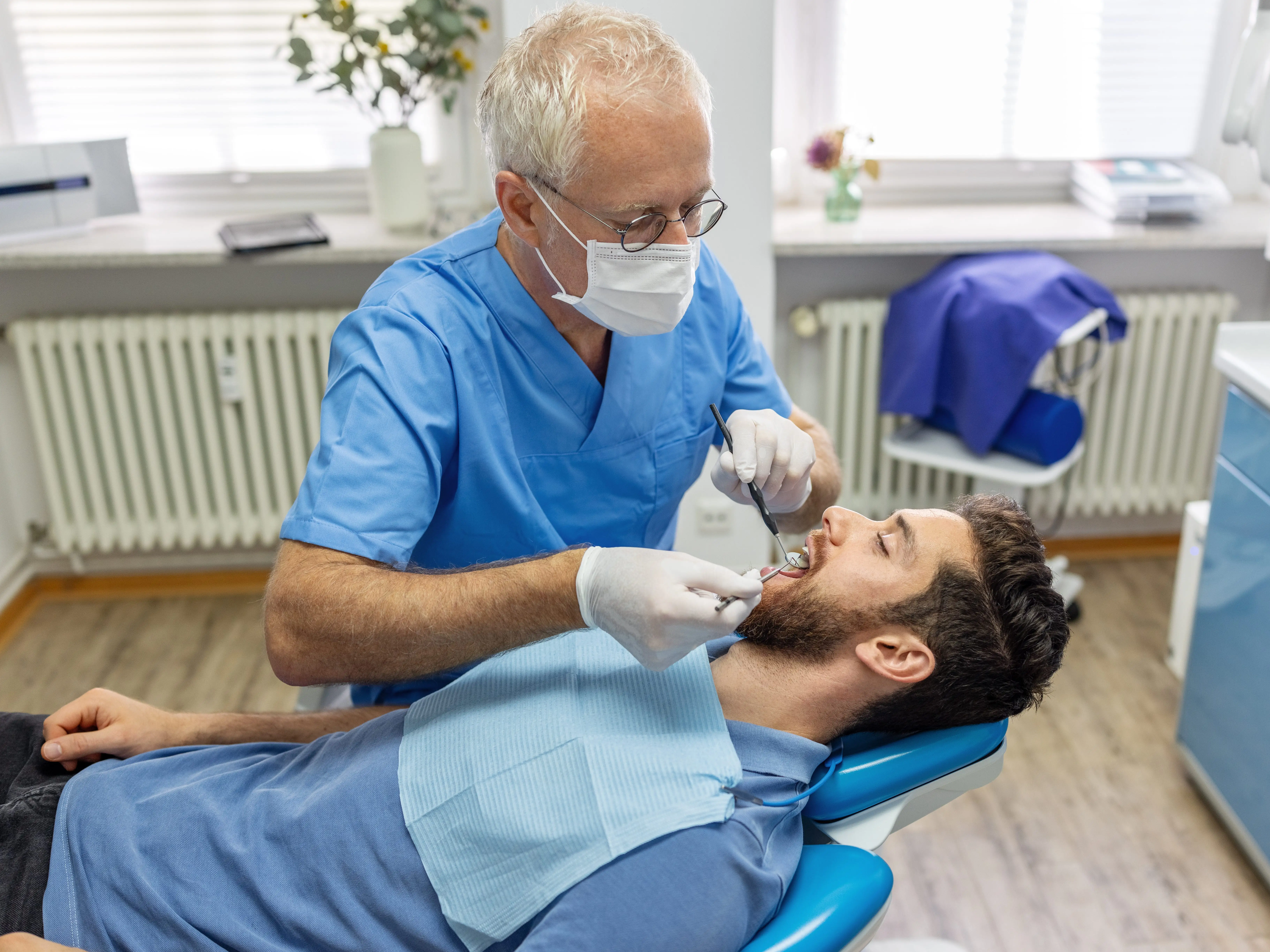 A dental professional in blue scrubs examines a reclined patient's teeth