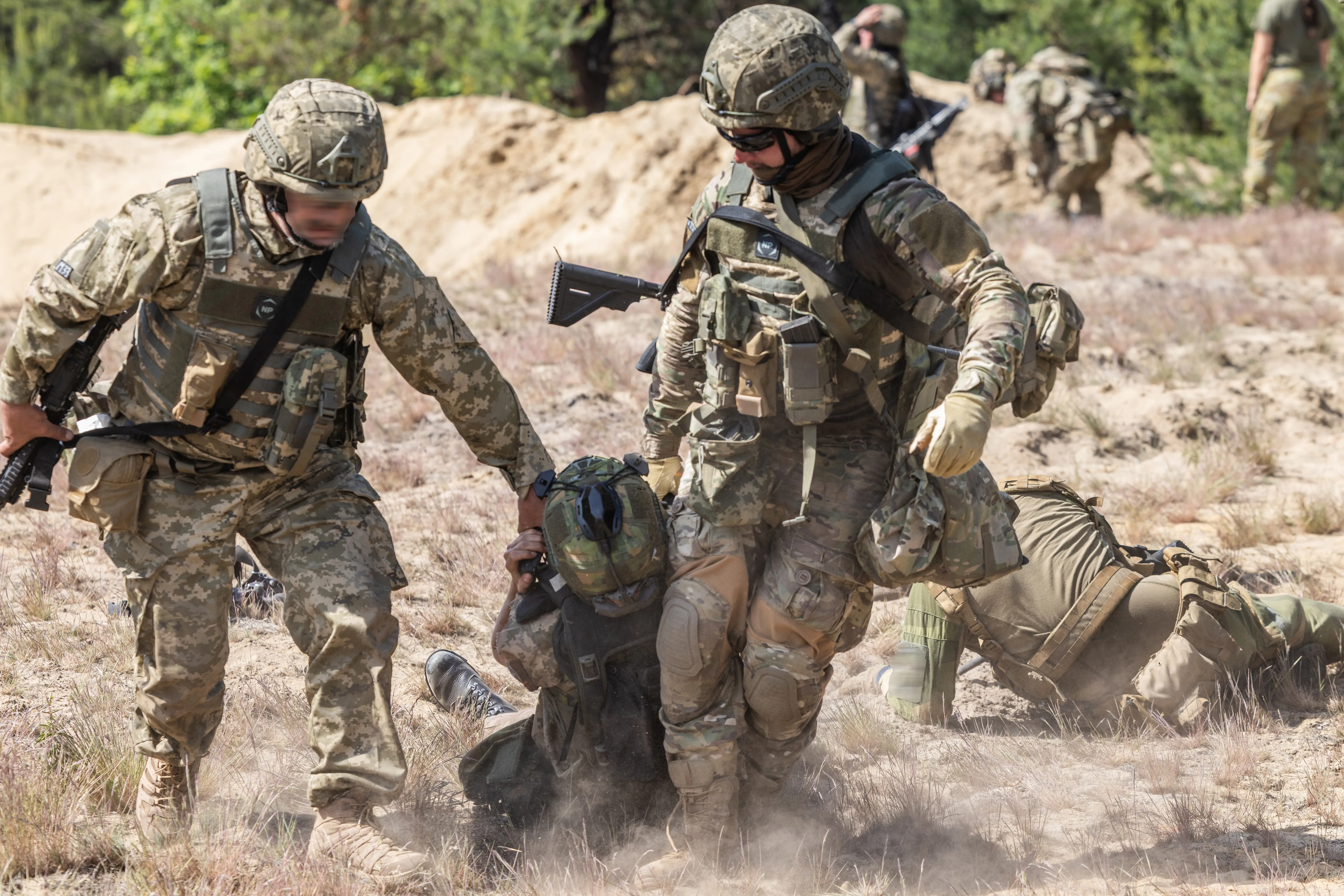Camouflaged military personnel assist a person on the ground during an outdoor field exercise with rifles and gear visible.