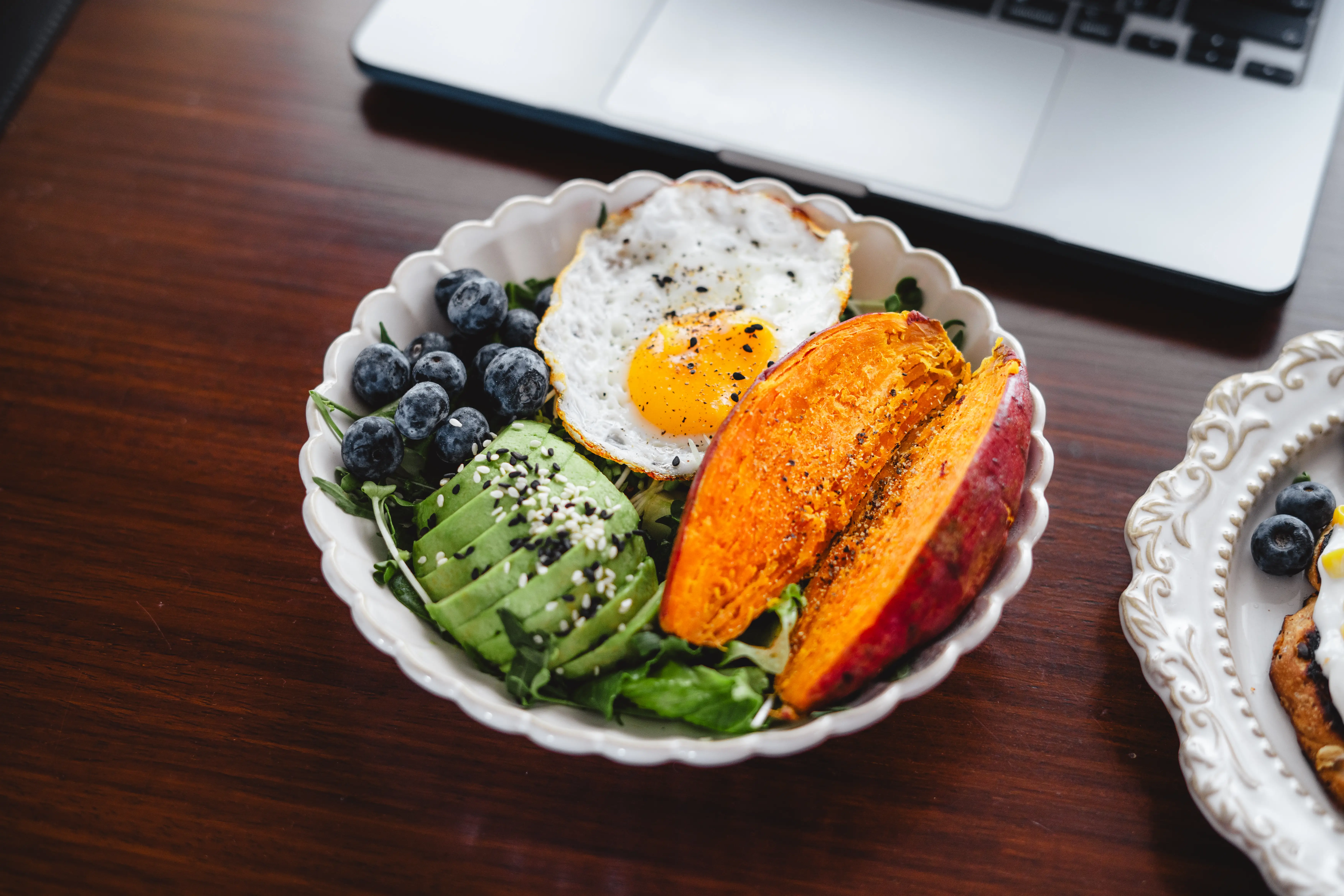 Bowl of avocado, blueberries, fried egg, sweet potato slices, and greens on a wooden table beside a laptop.
