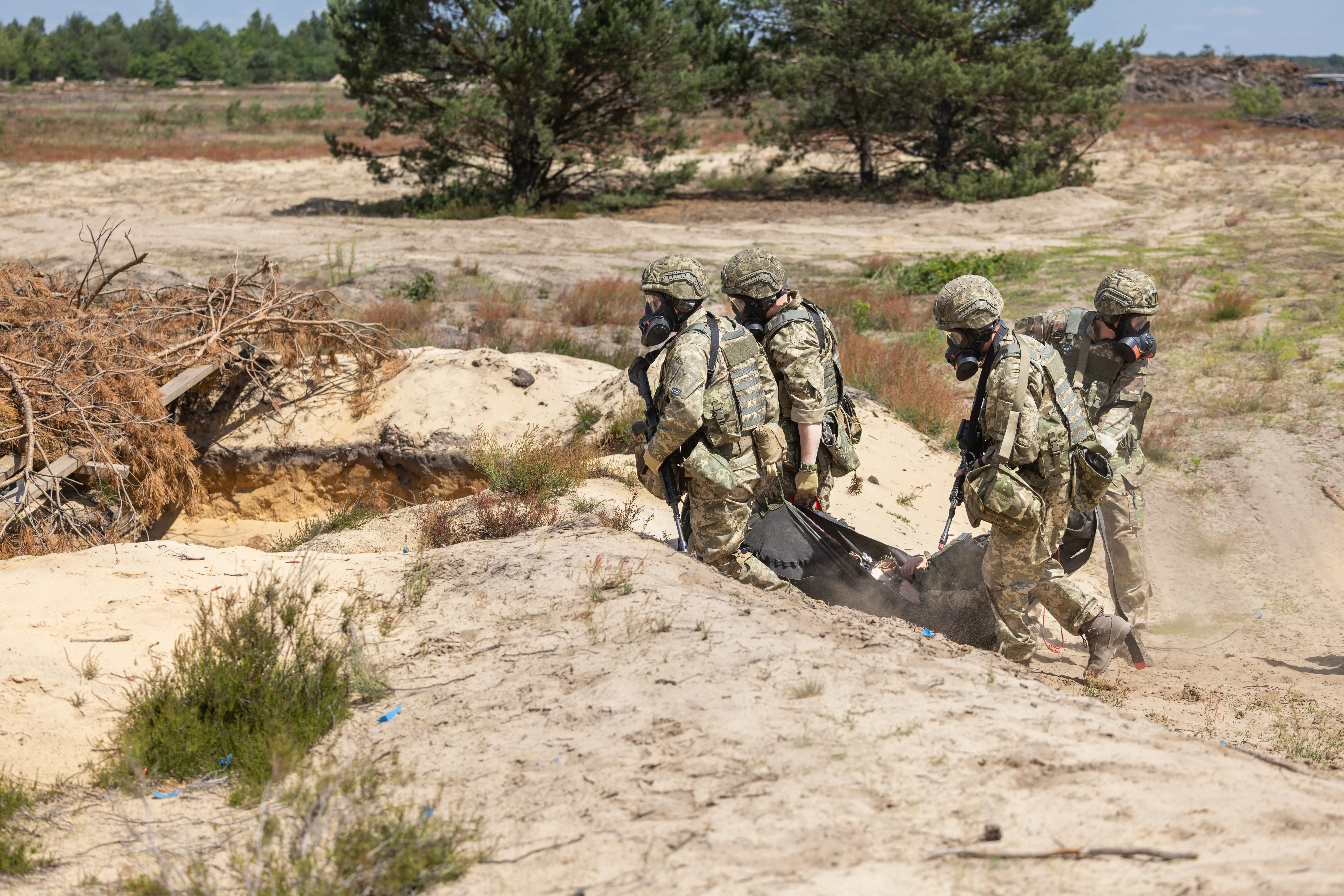 Camouflaged personnel wearing gas masks carry a stretcher across sandy terrain during an outdoor exercise.
