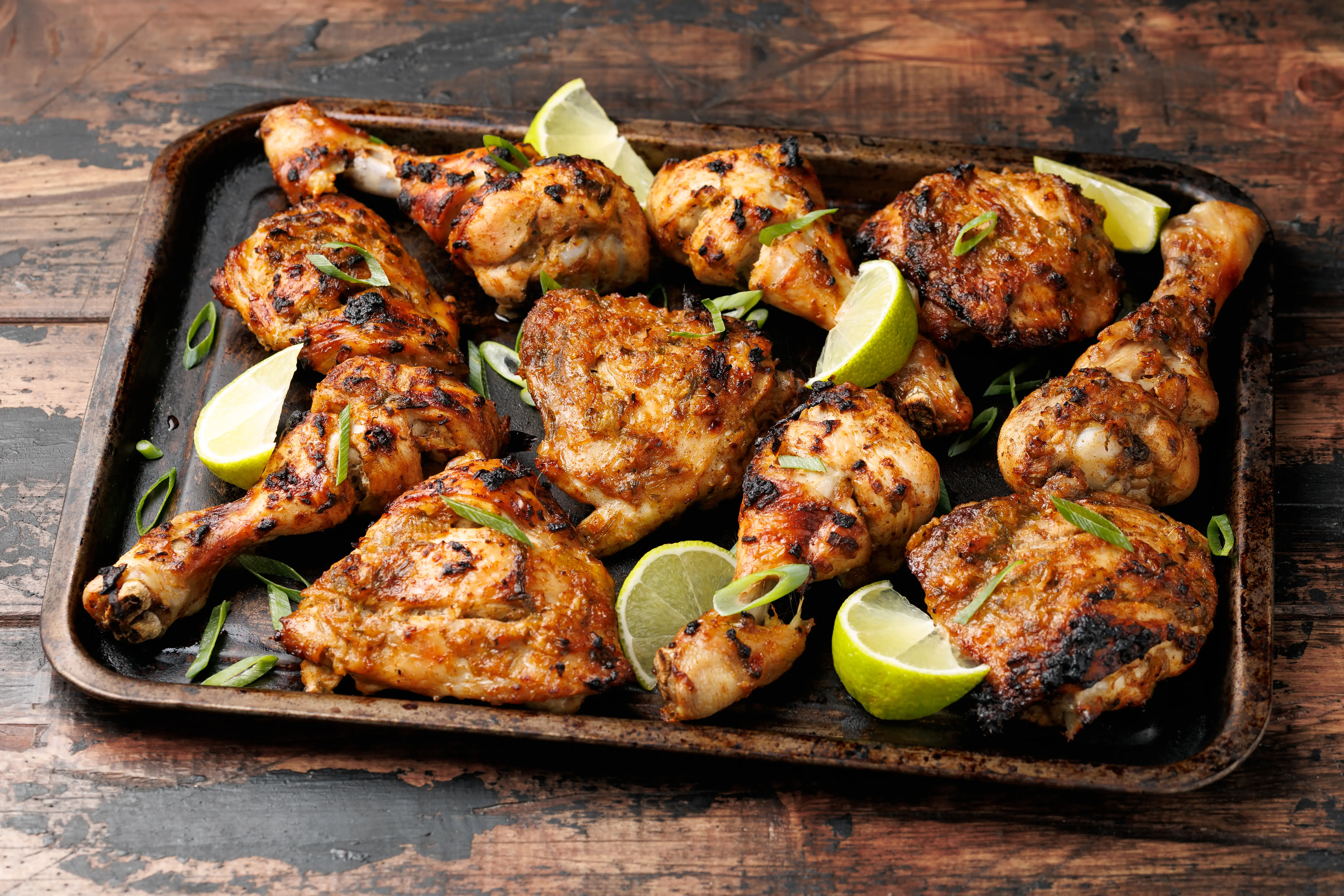 Grilled chicken pieces with lime wedges and sliced green onions on a dark tray over a wooden table.