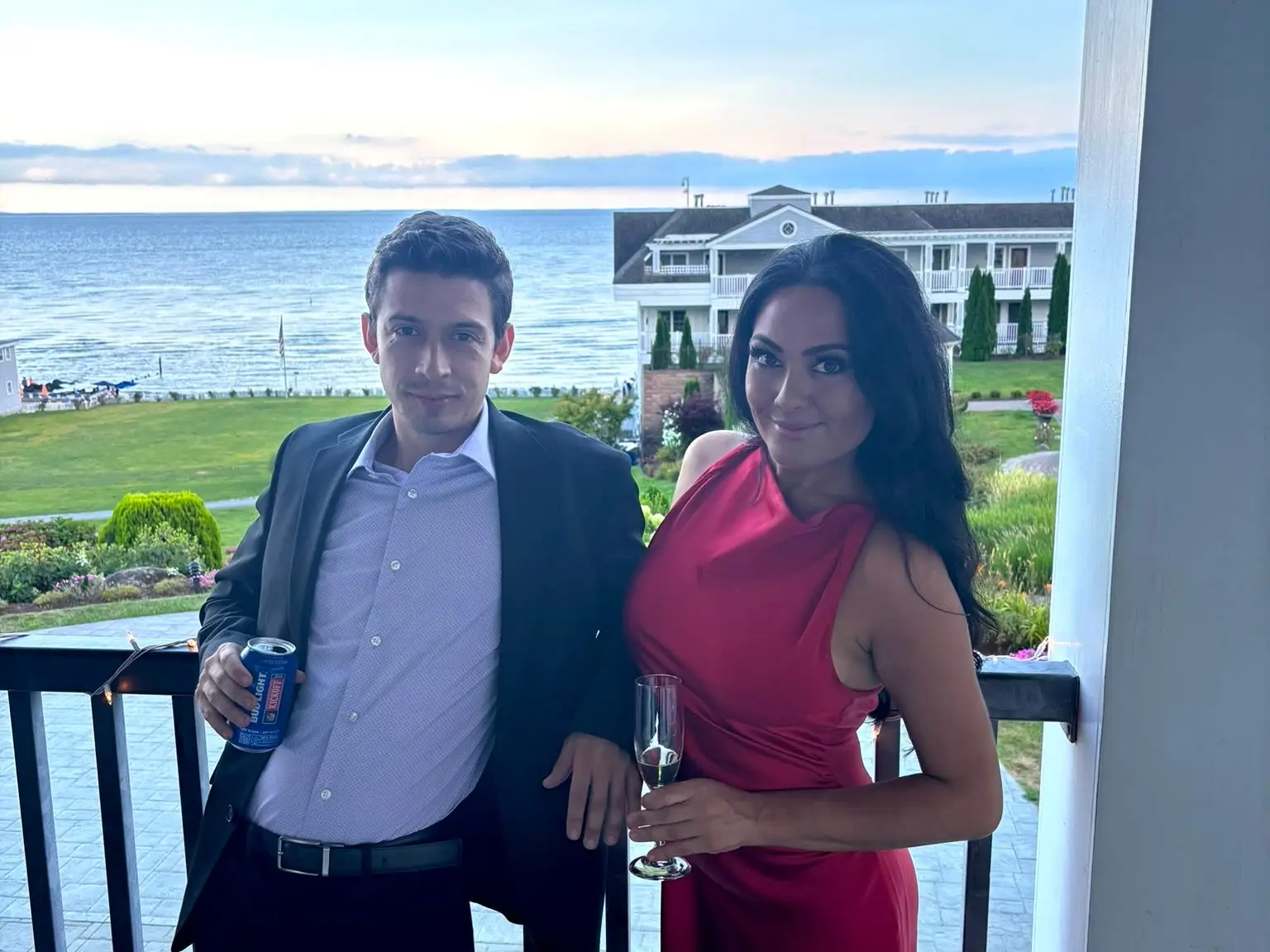 The writer and her partner pose with drinks on a balcony overlooking a waterfront lawn and buildings at sunset.