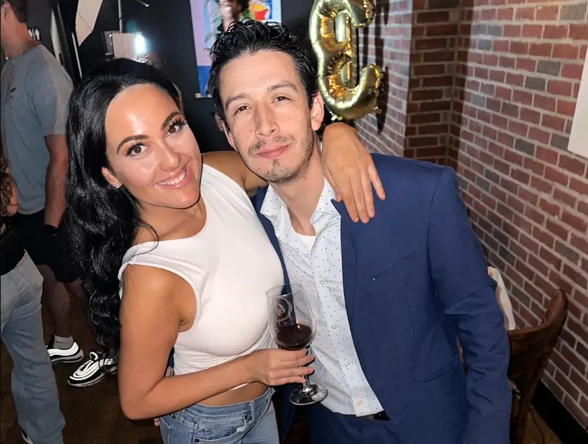 The writer holding a glass of wine and standing next to her partner, dressed in a suit, with a birthday balloon in the background.