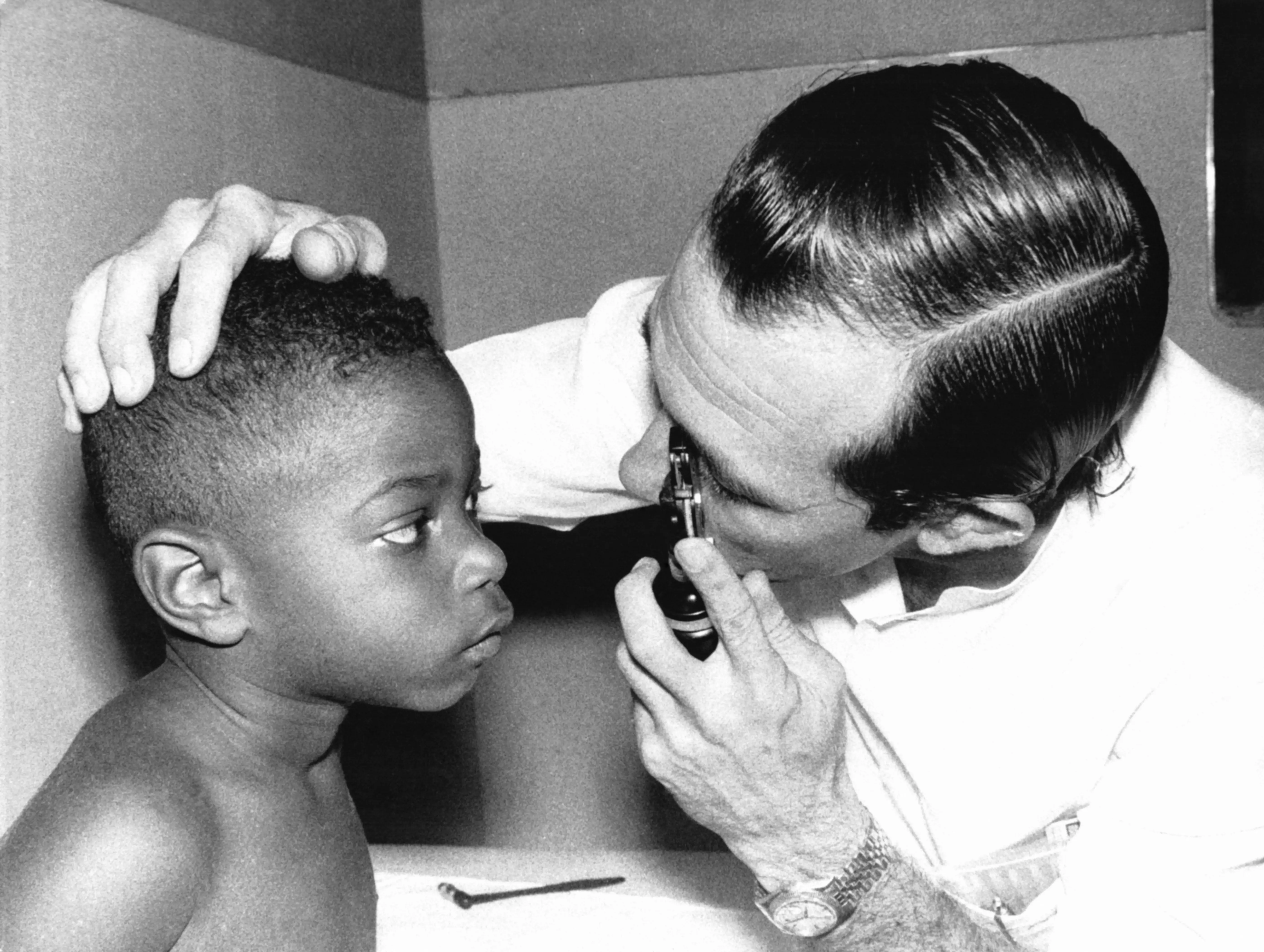 A clinician examines a patient's ear with an otoscope in a black-and-white medical scene.