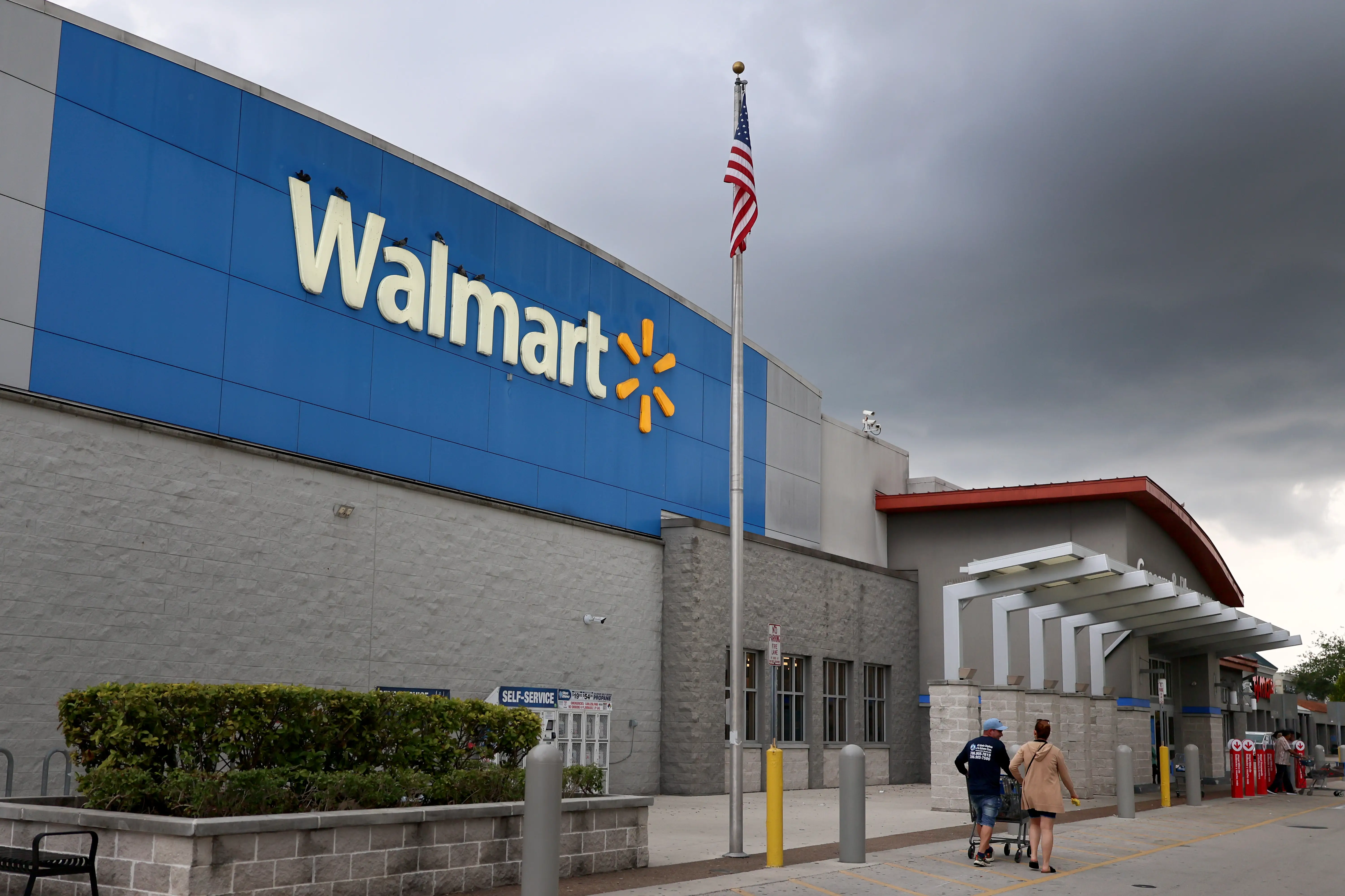 Walmart storefront with an American flag and shoppers near the entrance under dark cloudy skies.