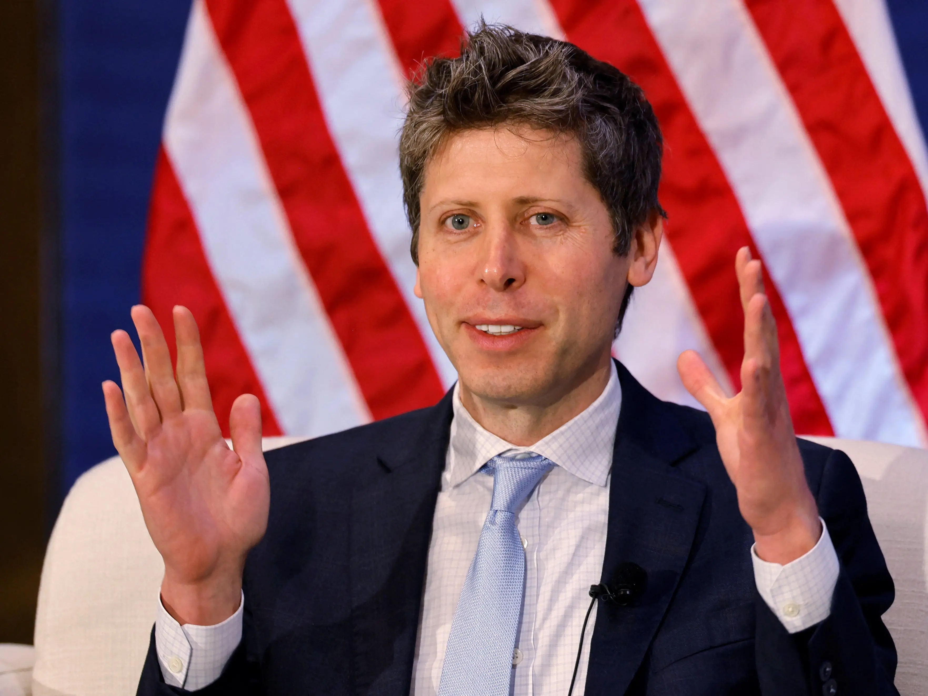 Sam Altman speaks with raised hands in front of a United States flag backdrop with a Reuters watermark.
