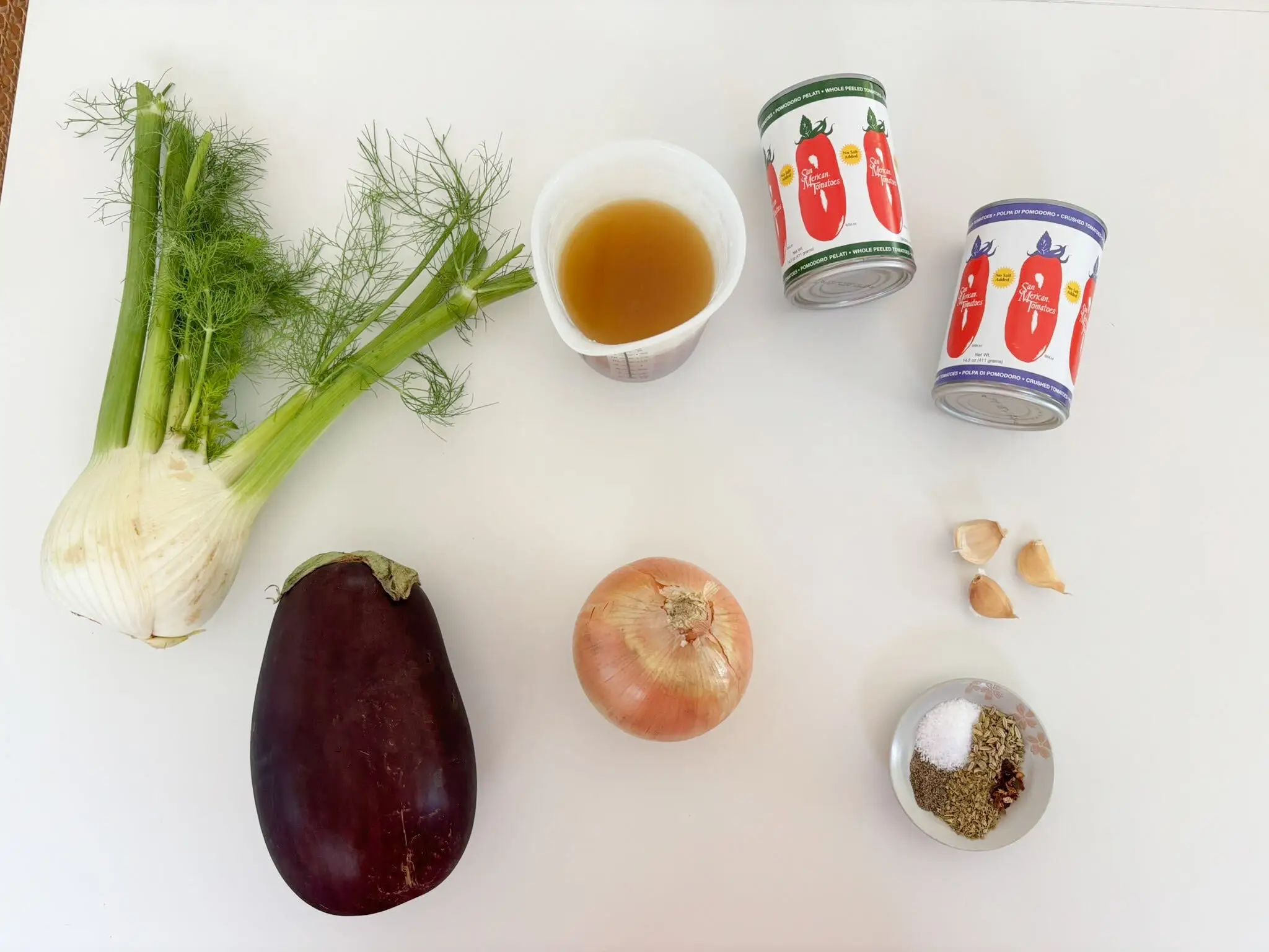 Overhead arrangement of fennel, eggplant, onion, garlic, spices, amber liquid, and tomato cans sitting on a white surface.