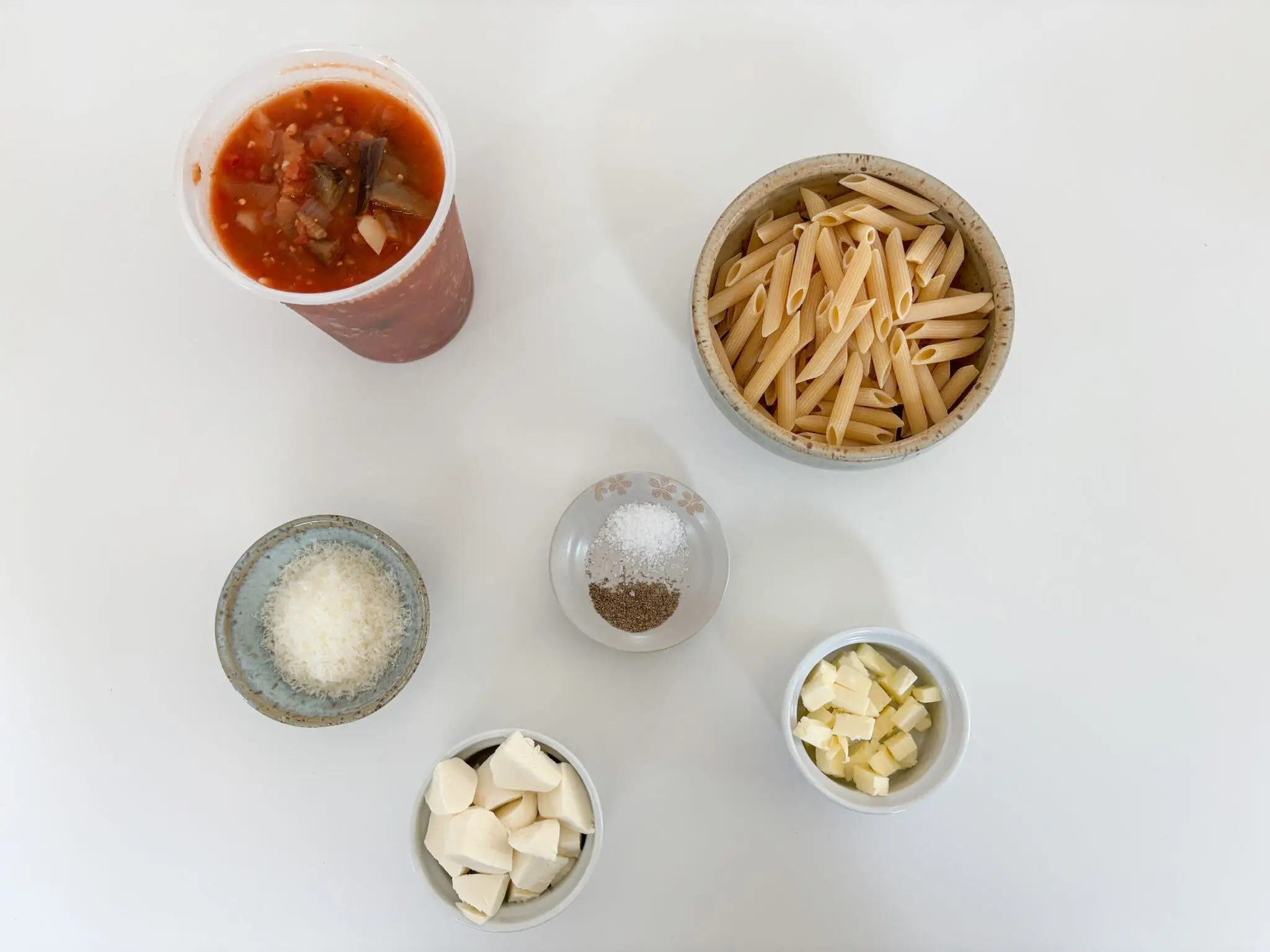 Overhead arrangement of pasta ingredients with penne, tomato-eggplant soup, grated cheese, seasonings, garlic, and diced butter on a white surface.