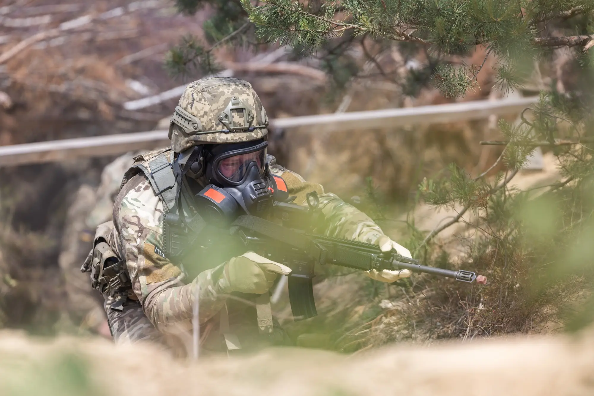 Camouflaged soldier wearing a gas mask aims a rifle from cover in a wooded outdoor training area.