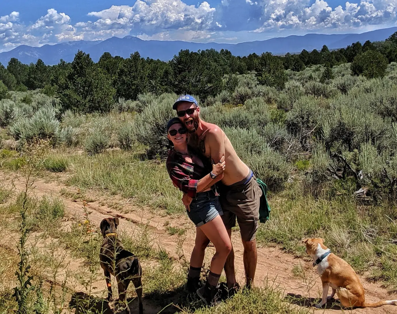 The writer hugging her husband in front of trees and woodland near their house, with two dogs next to them.