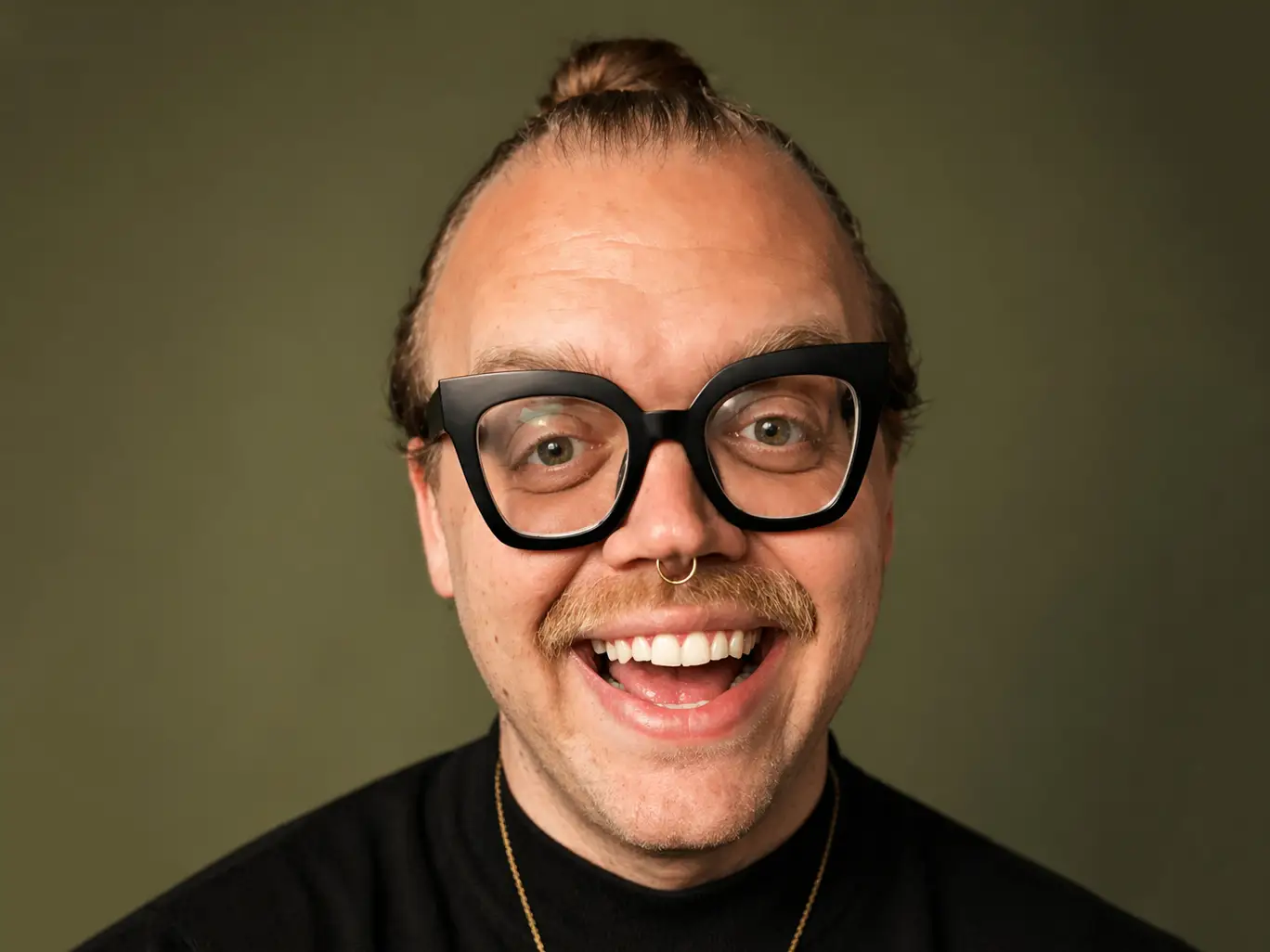 Smiling person wearing black glasses, a nose ring, black turtleneck, and gold heart pendant against an olive background.