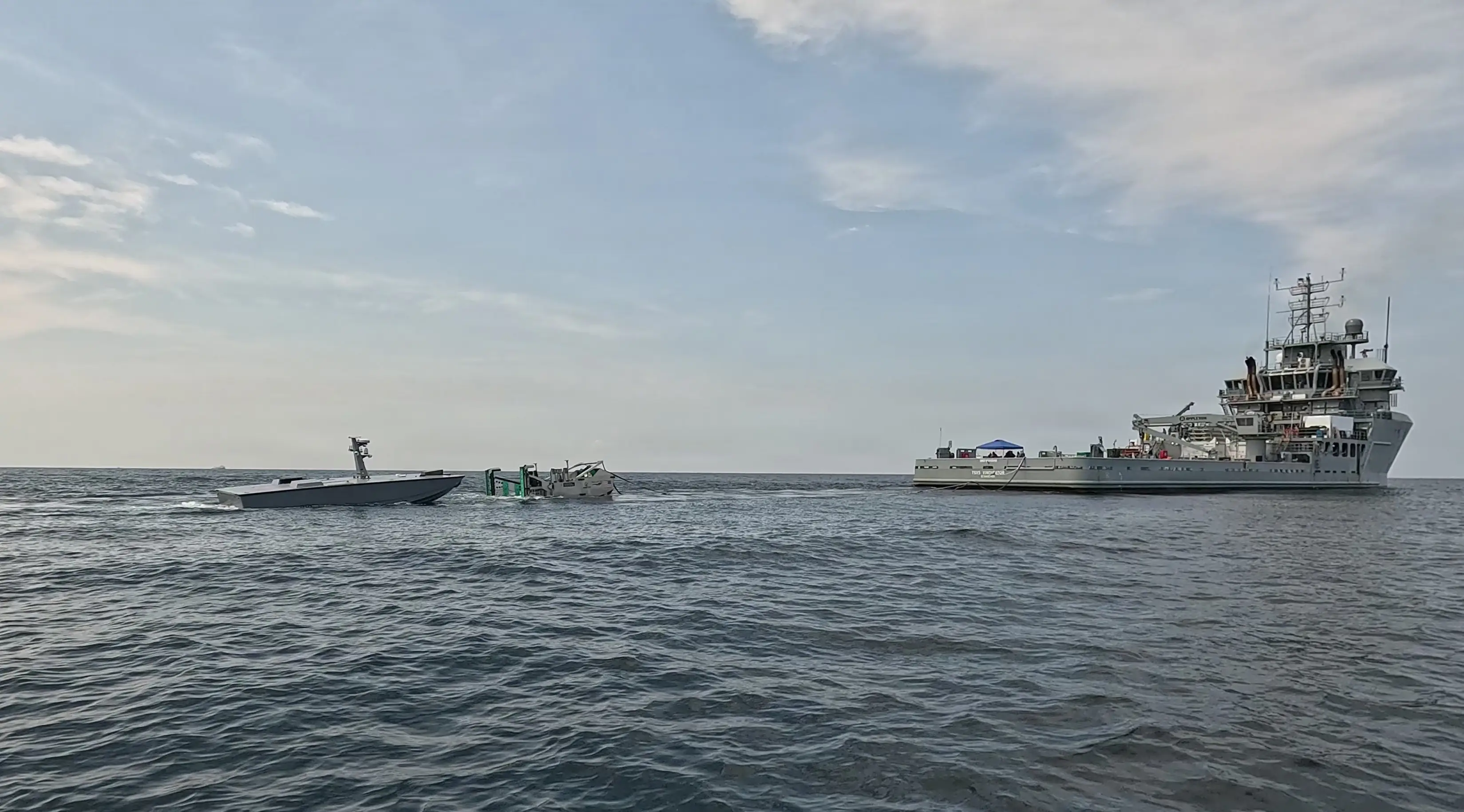 A naval drone and robotic refueling system float in blue ocean water near a US Navy vessel.