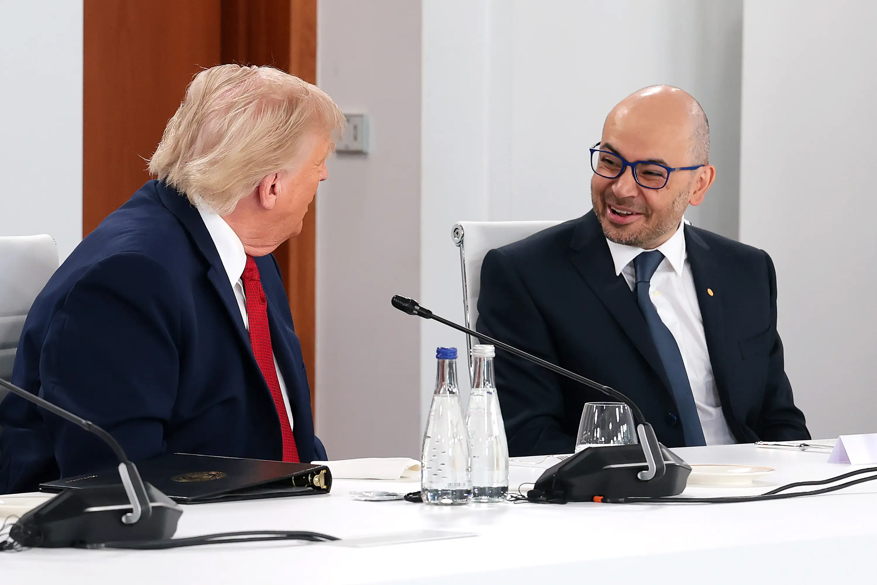 Donald Trump sits at a conference table speaking with a suited official beside microphones and water bottles.