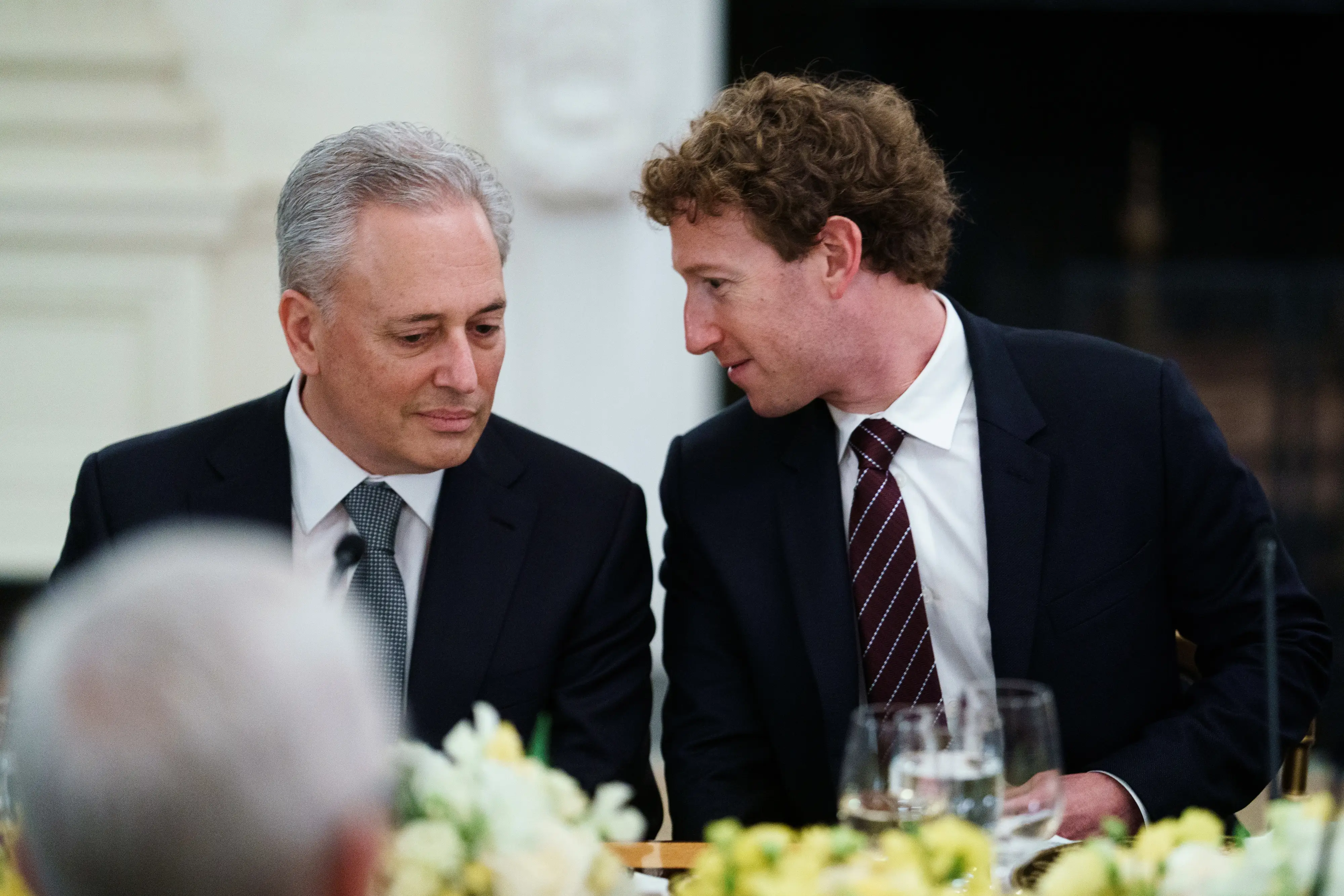 Mark Zuckerberg speaks with David Sacks at a formal dining table with glasses and floral centerpieces.