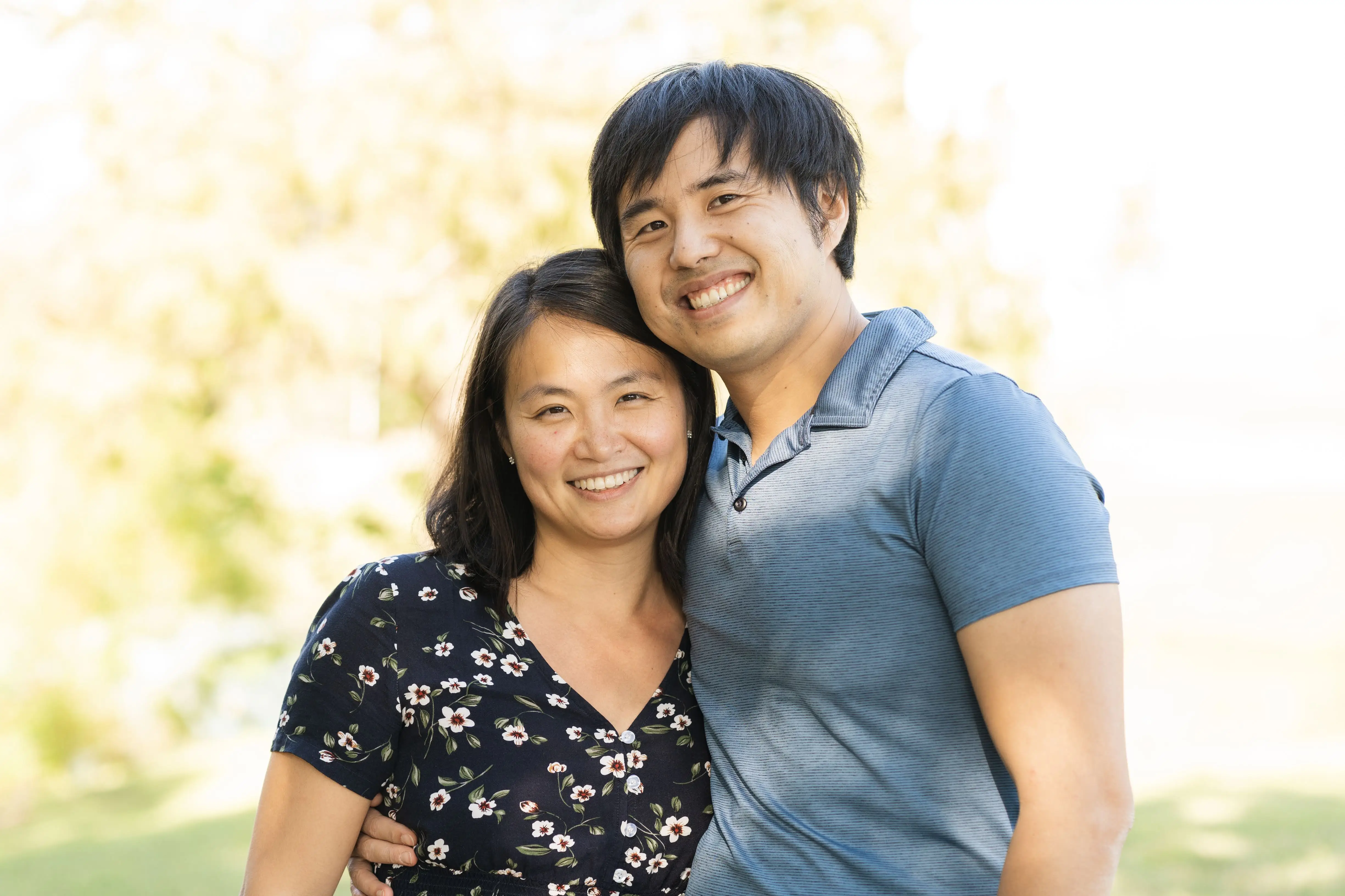 Two smiling people stand close together with arms around each other in a sunlit outdoor setting.