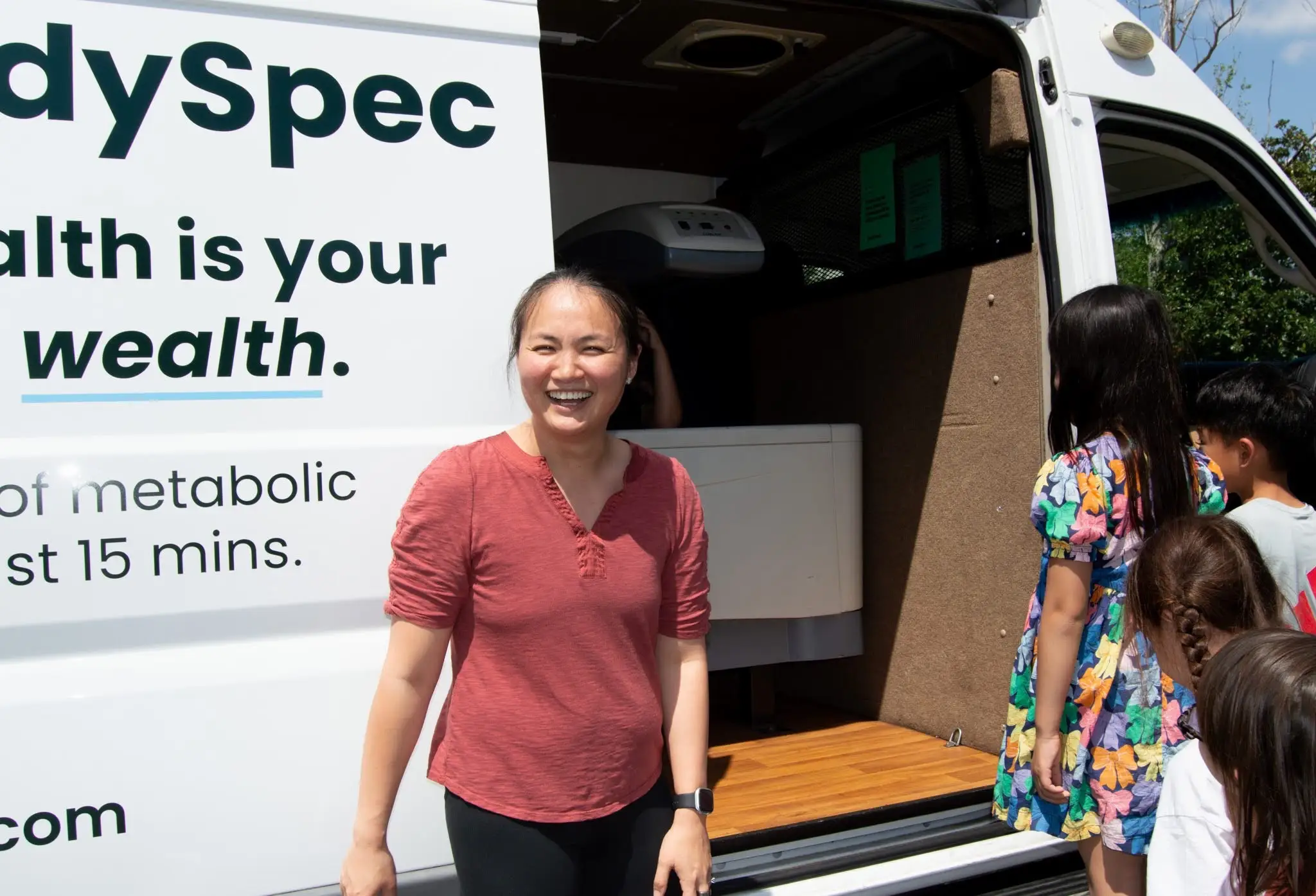A smiling person stands beside an open mobile screening van as several people wait near the entrance.