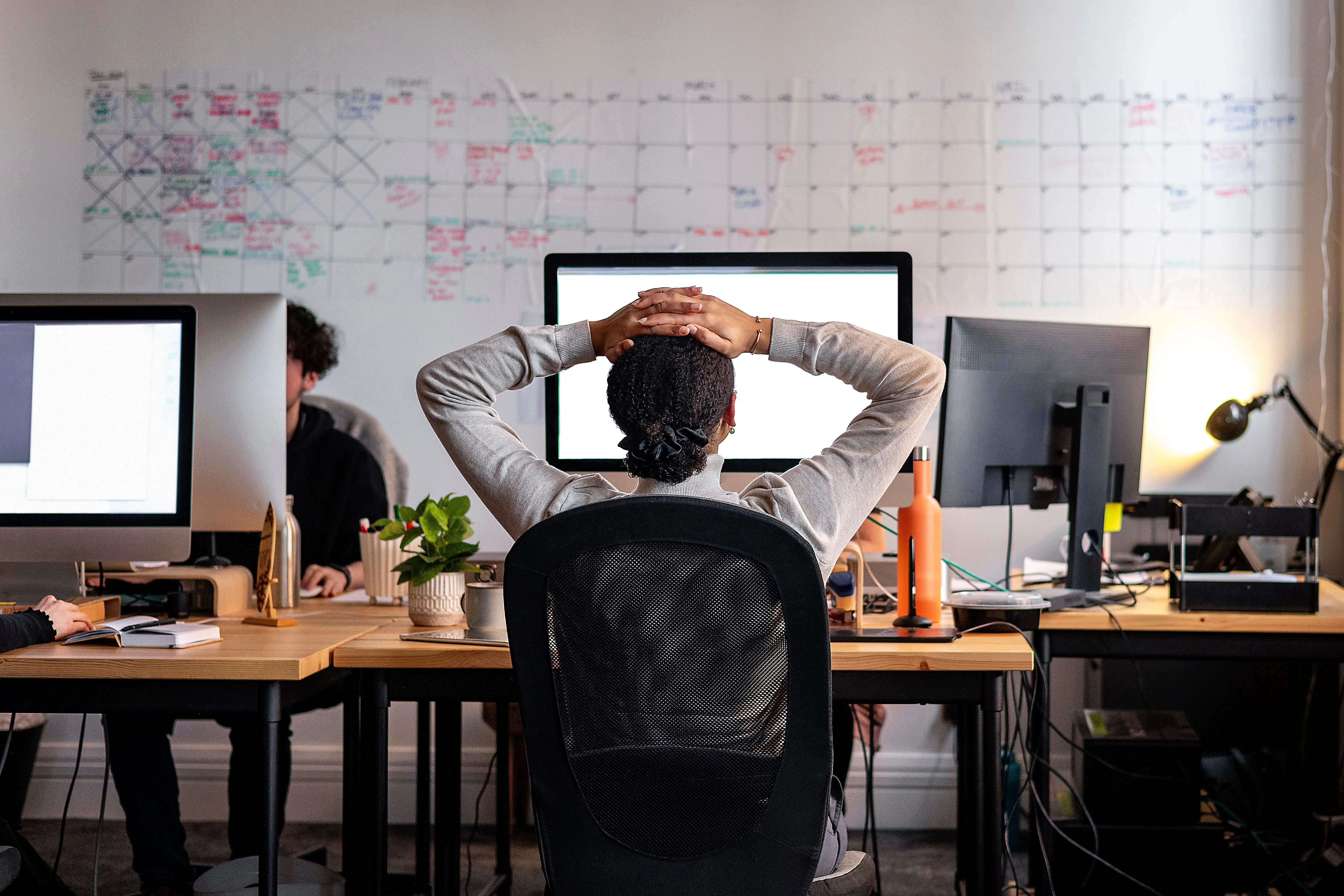 Rear view wide shot of a woman sitting working at a computer in an office. She has her hands behind her head as she stretches, looking at the screen.
