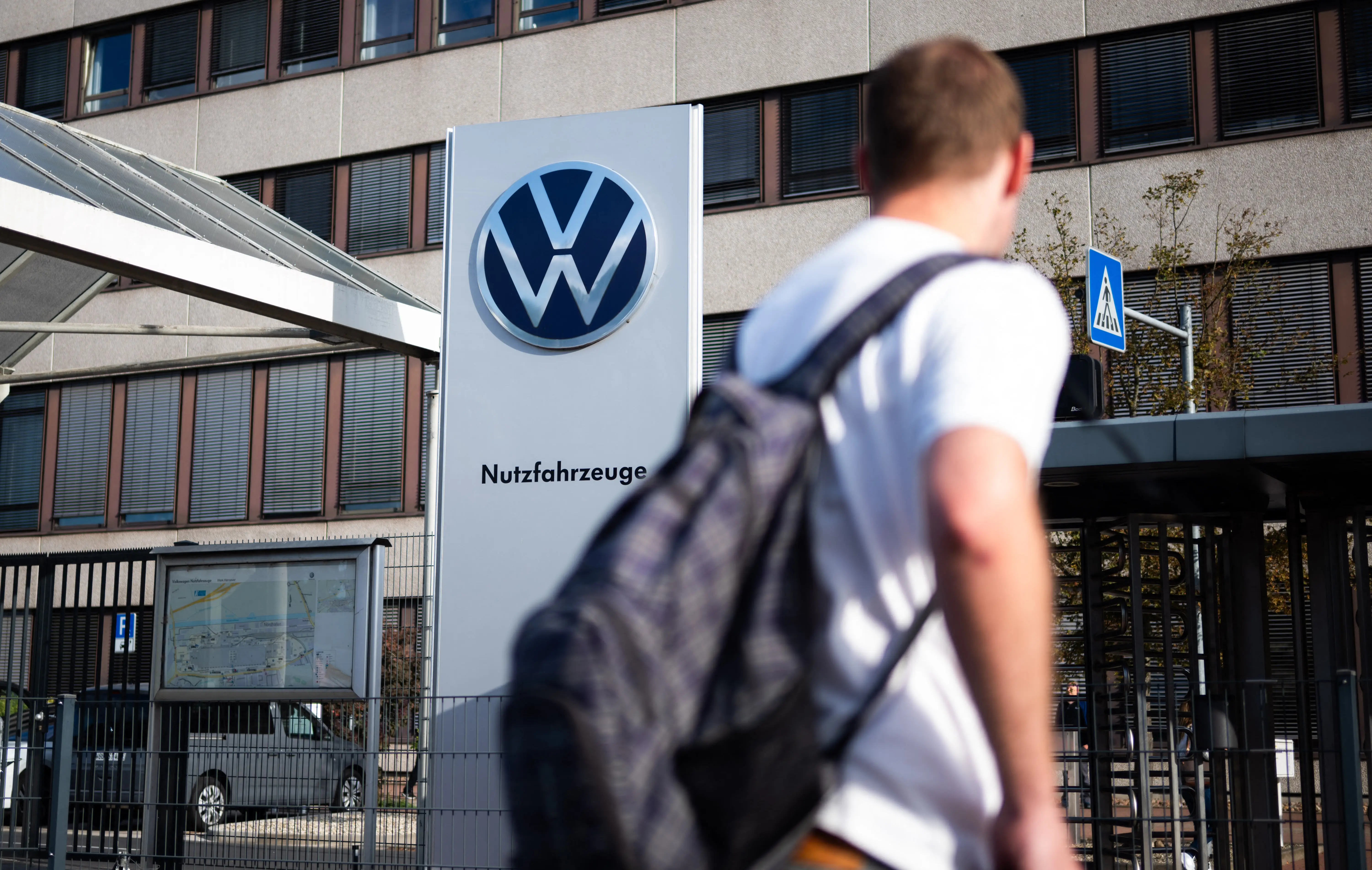 A person wearing a white shirt and a backpack walks past a Volkswagen sign.